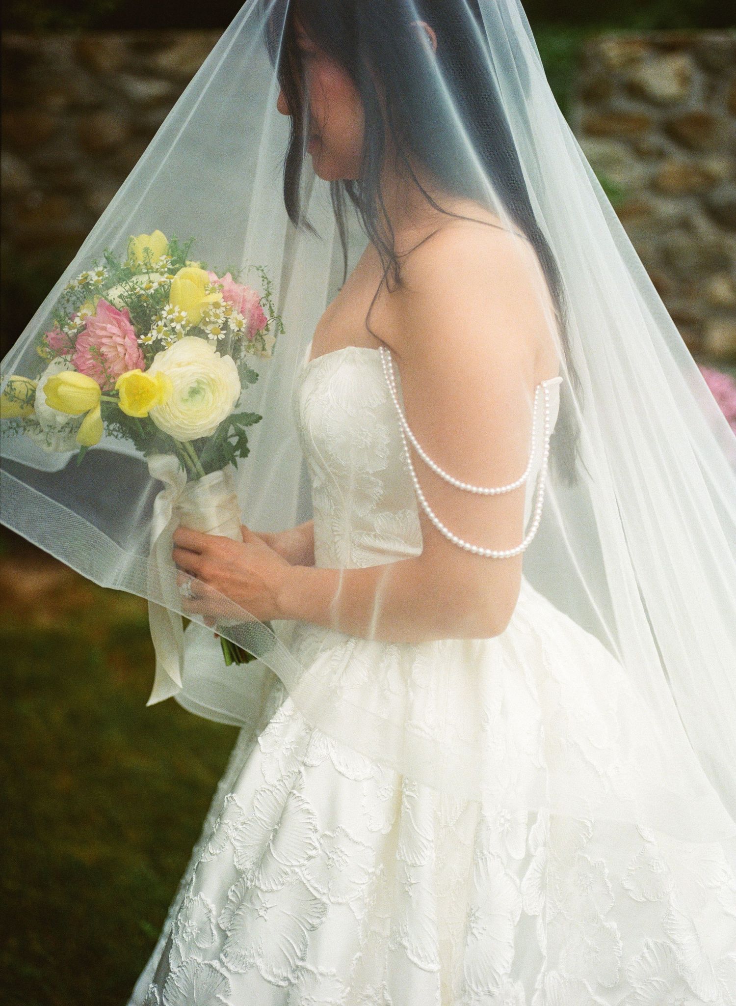 Bride in strapless lace gown with pearl drape back, holding yellow and pink bouquet under sheer wedding veil.
