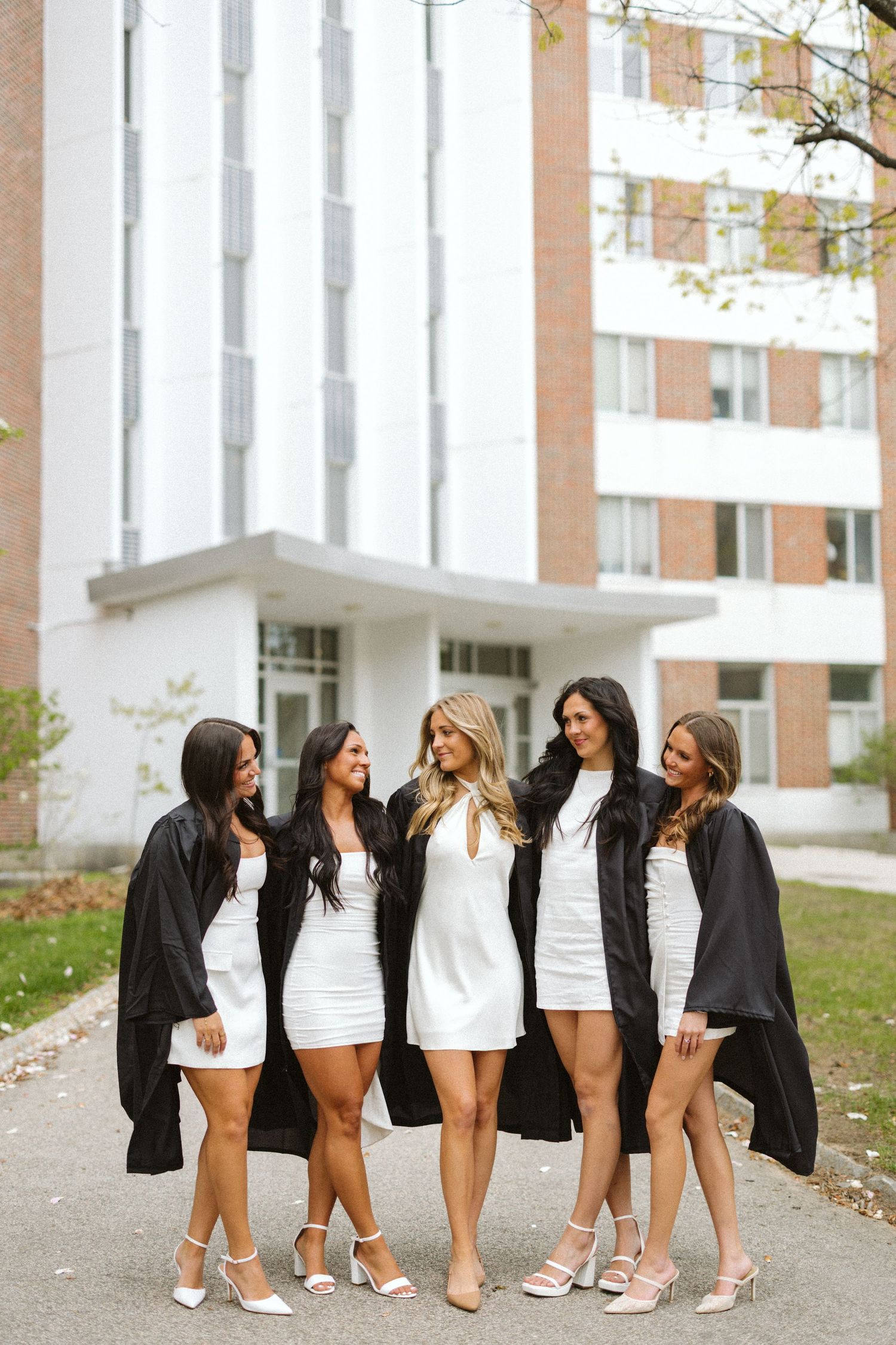 Five young women in white dresses and black graduation gowns smiling together outside a university building.