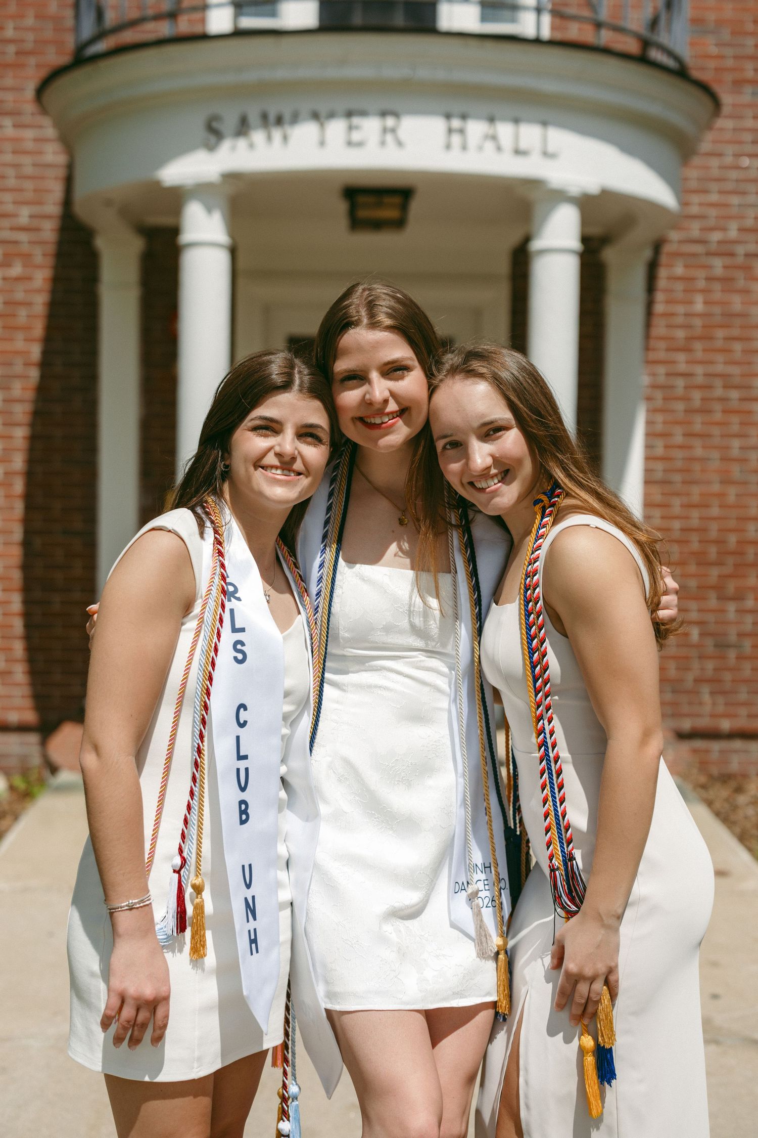 Three smiling graduates in white dresses pose with honor cords in front of Sawyer Hall on graduation day.