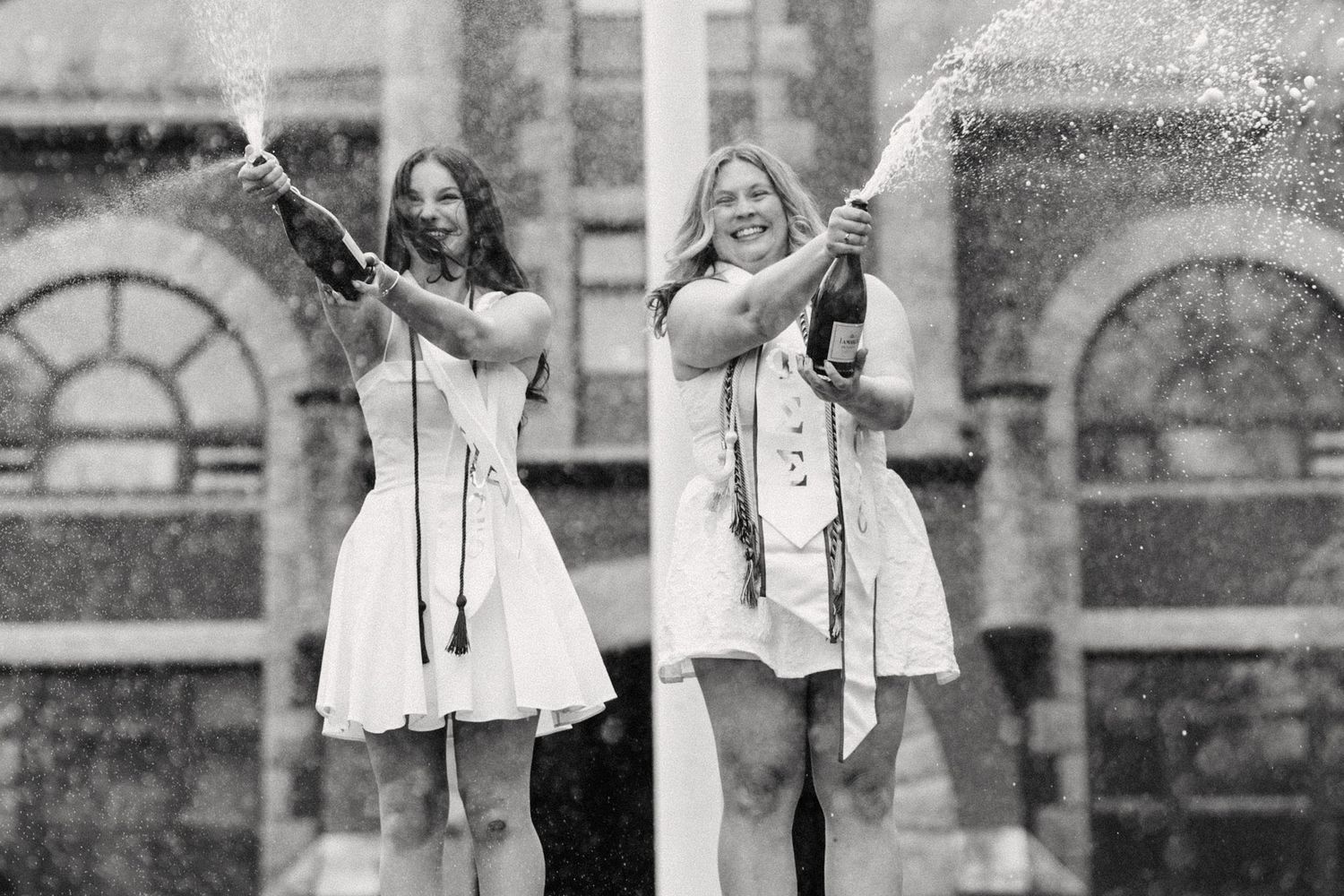 Two women in white dresses celebrate with champagne spray in a black and white photo outdoors.