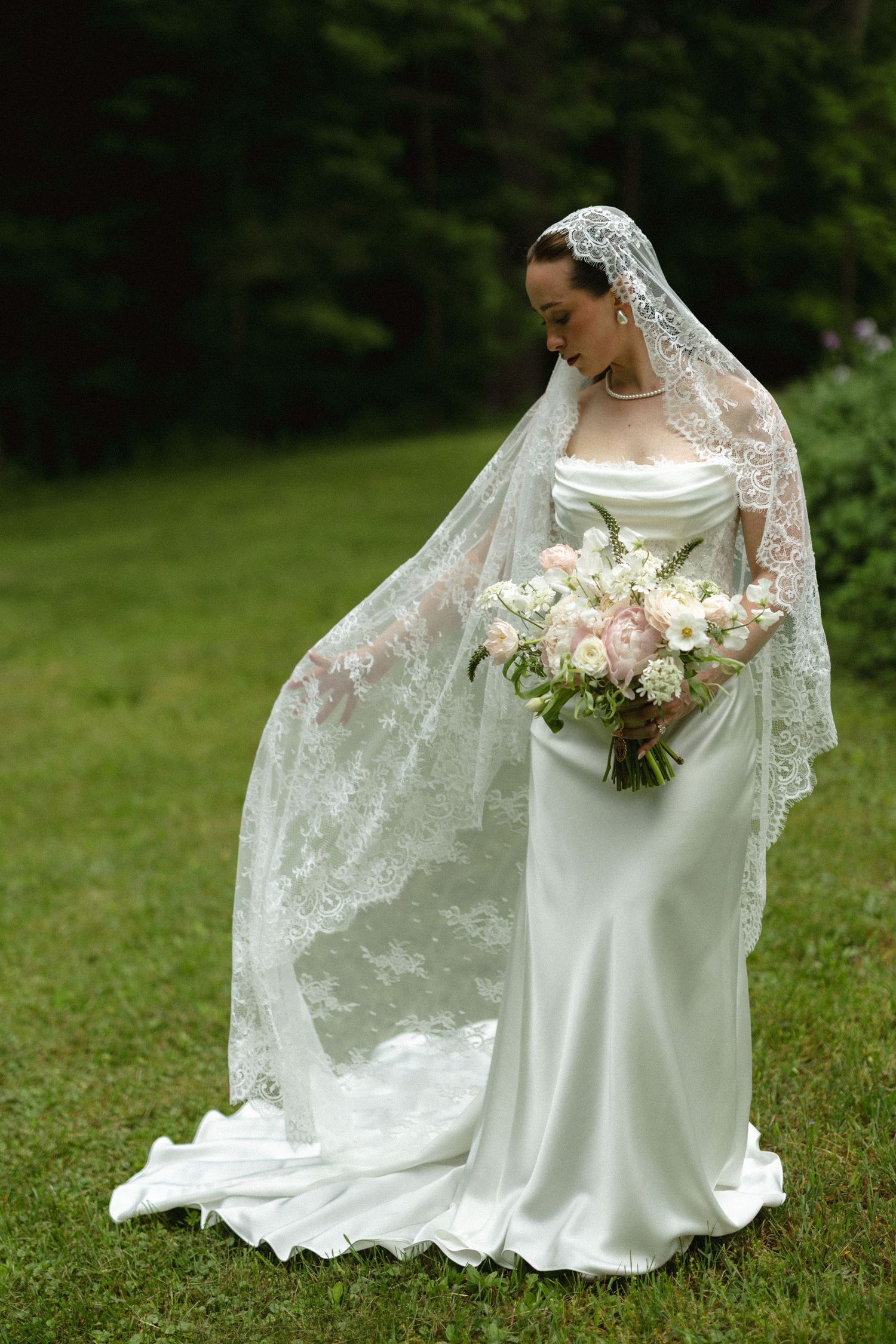 Elegant bride in white satin gown and lace mantilla veil holding pink and white bouquet outdoors.