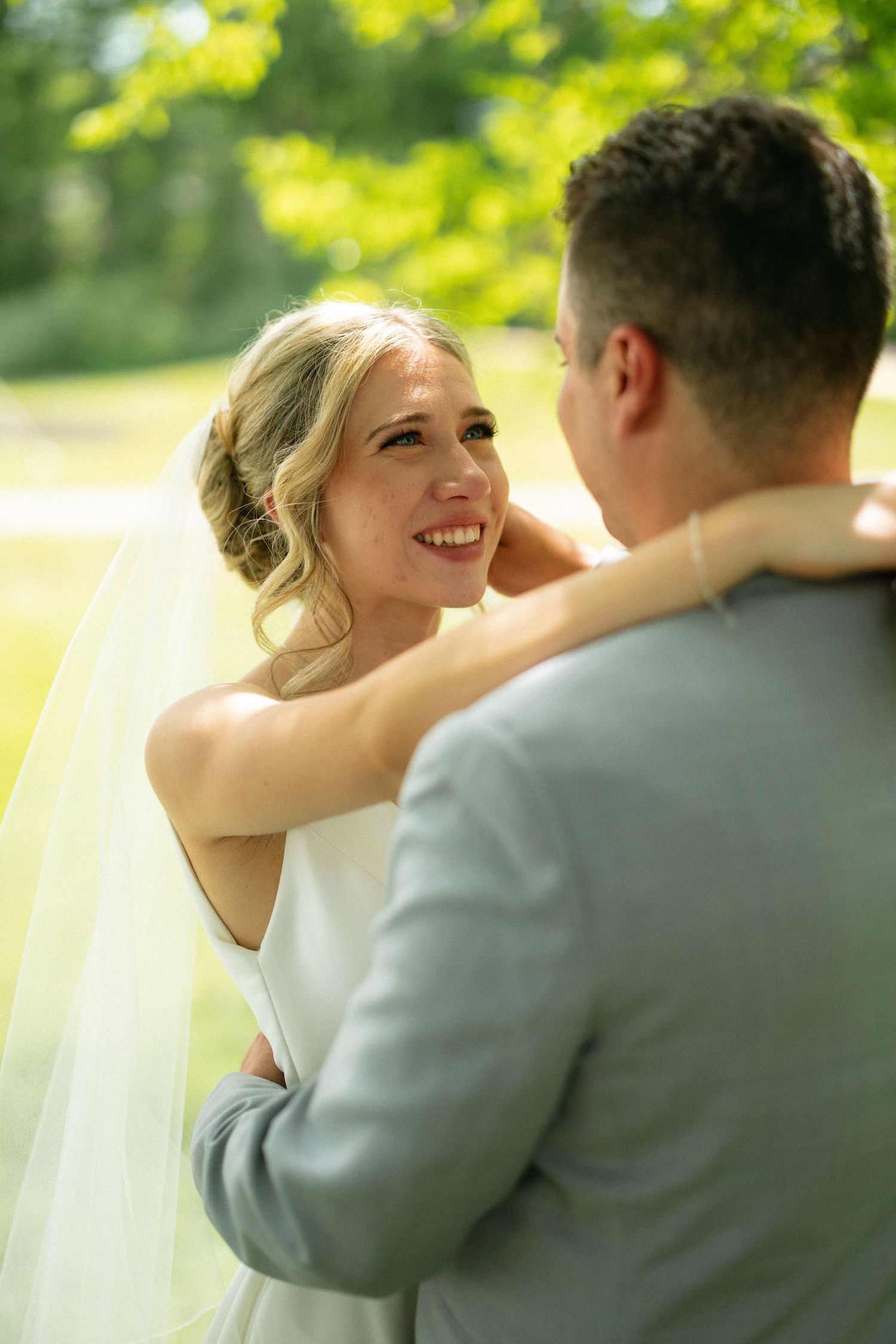 Smiling bride in white dress and veil embraces groom in gray suit outdoors in lush green garden setting.