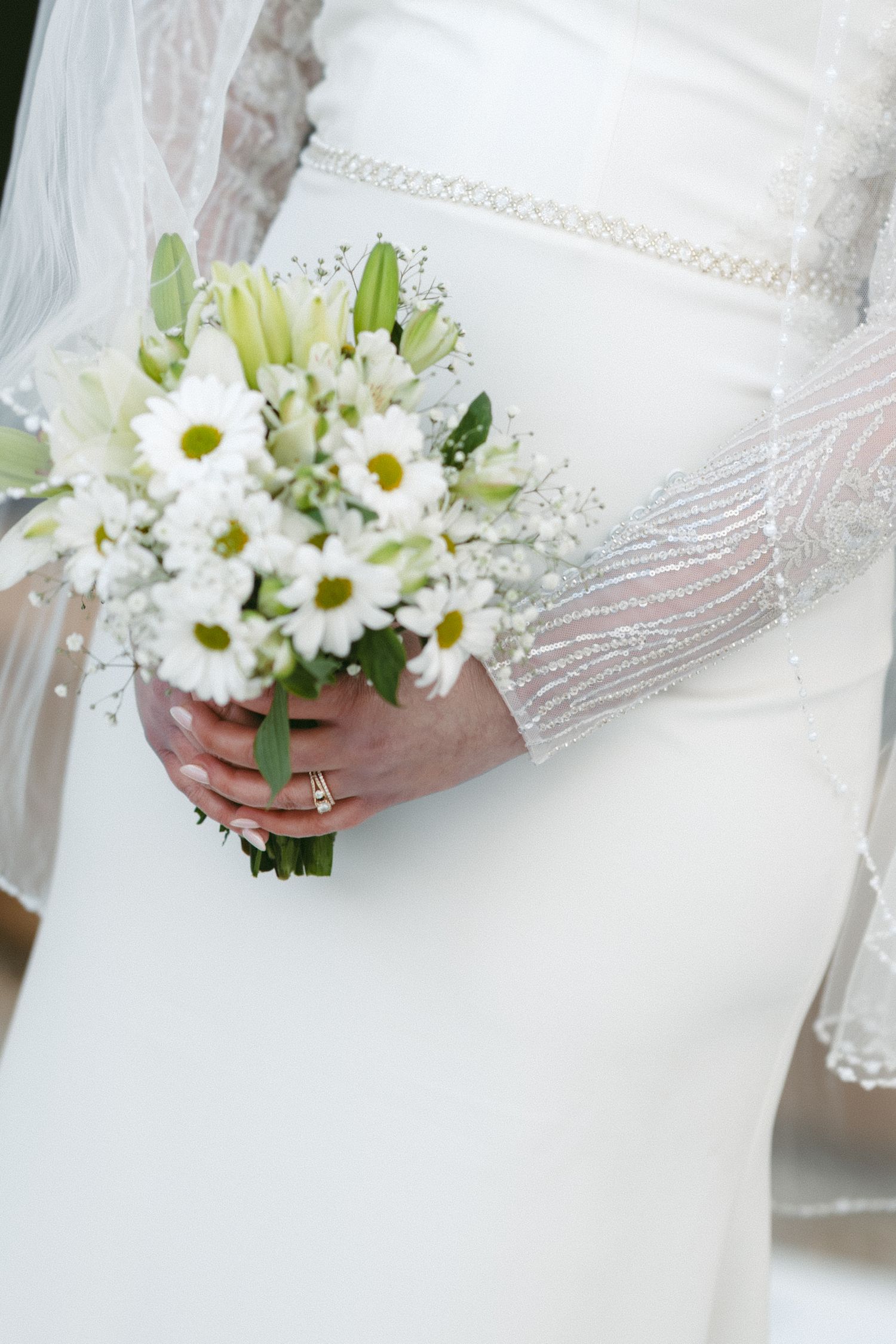 A delicate white bridal bouquet of daisies held against a flowing white wedding dress.