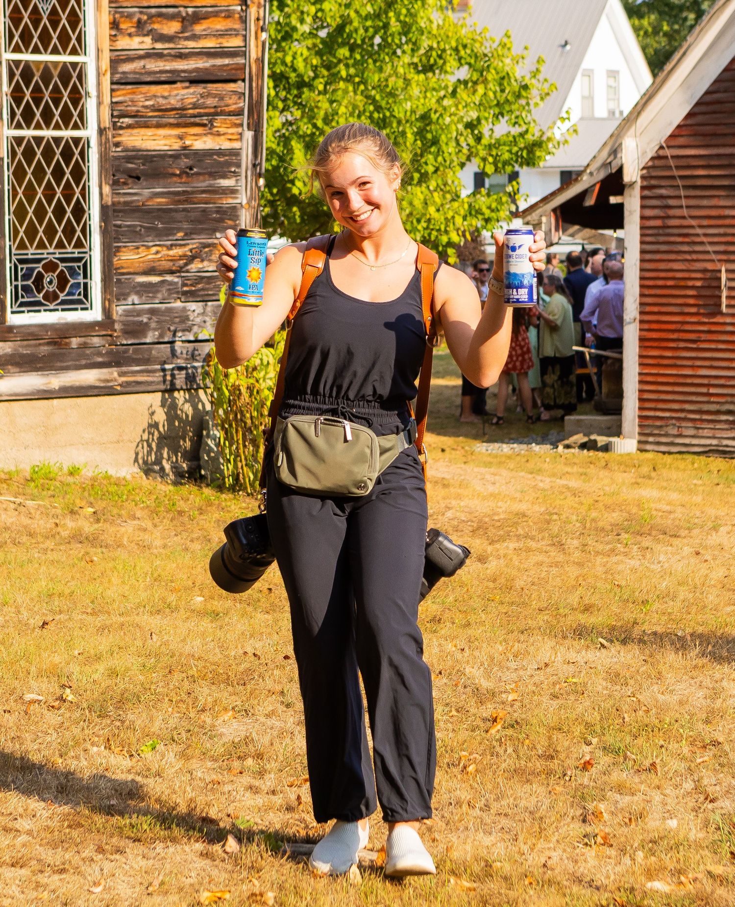 Someone in casual athletic wear holds up a drink while standing on grass near rustic wooden buildings on a sunny day.