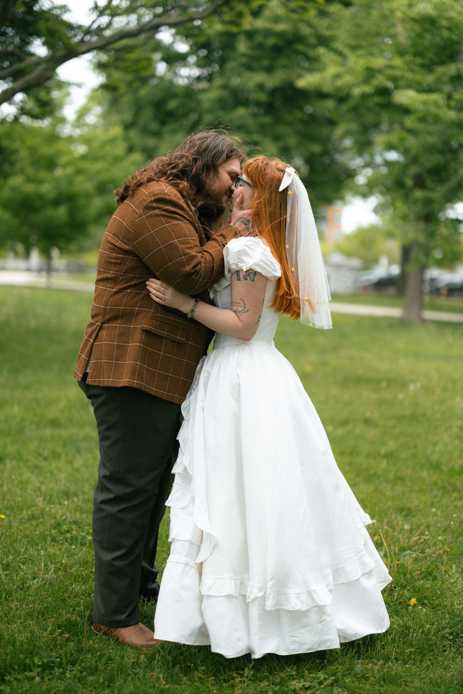 A bride and groom share a romantic kiss outdoors on green grass, she in a white ruffled gown and veil, he in a brown plaid jacket.