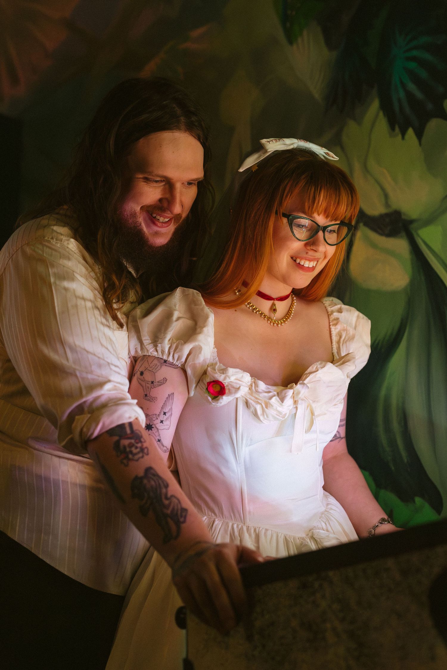 Tattooed man and redhead woman in white dress smile together against a tropical mural backdrop.