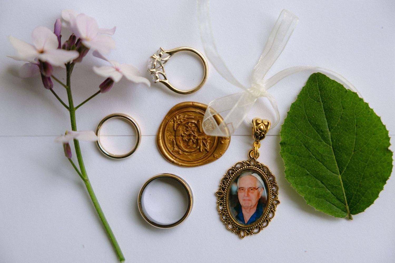 Gold wedding rings, diamond ring, wax seal, memorial locket with portrait, flower, and green leaf on white background.