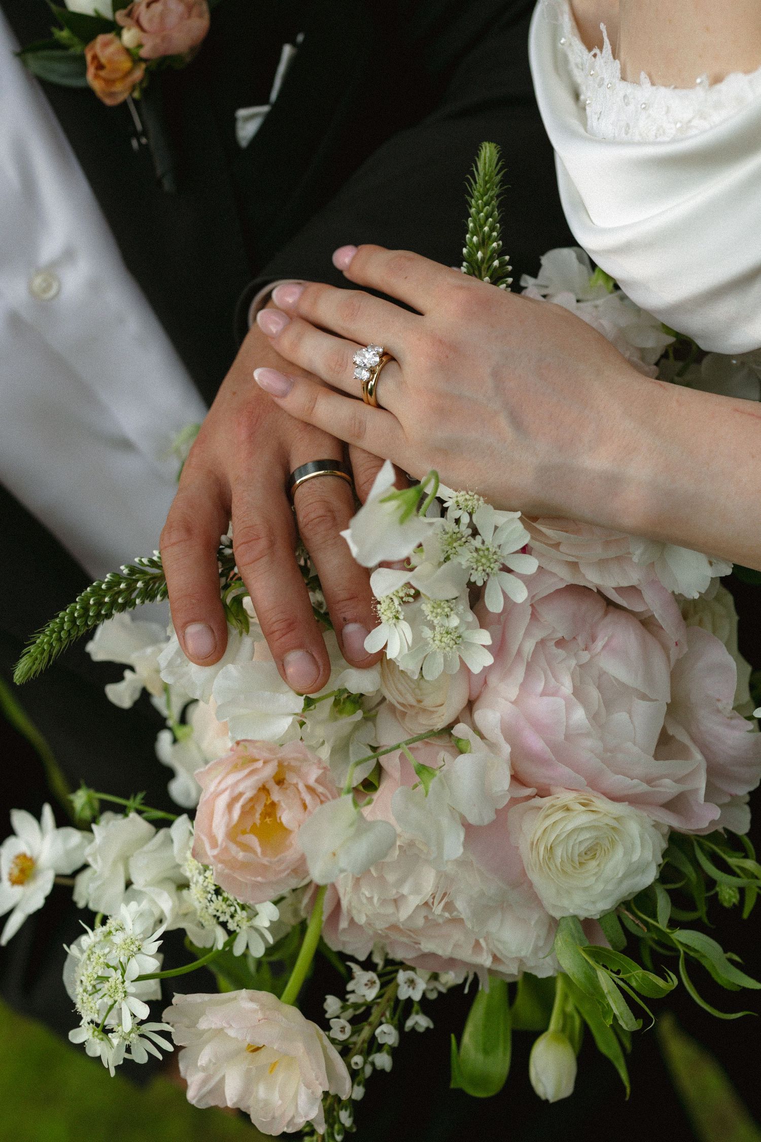 Bride and groom hands with wedding rings resting on a bouquet of pink peonies and white flowers.