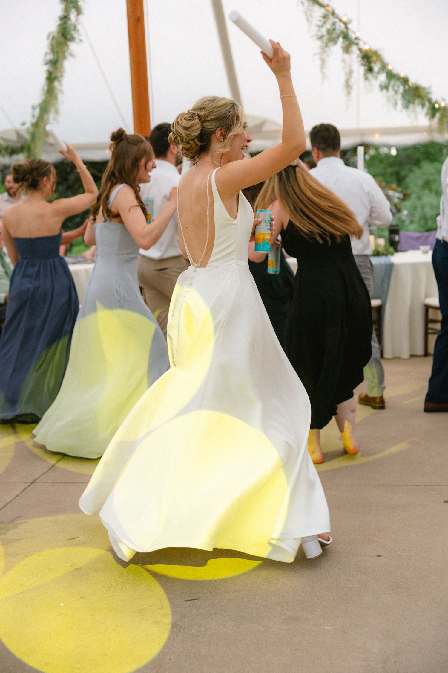 Bride twirling in white backless gown on dance floor surrounded by guests at outdoor tent wedding reception.