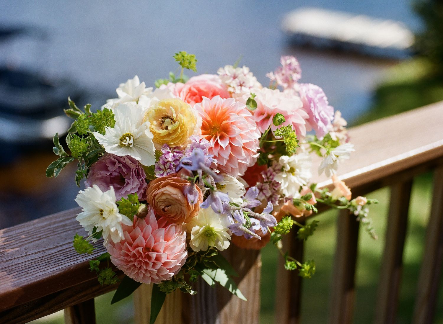 Lush bridal bouquet of pink dahlias, peach ranunculus, and white blooms resting on a wooden railing outdoors.