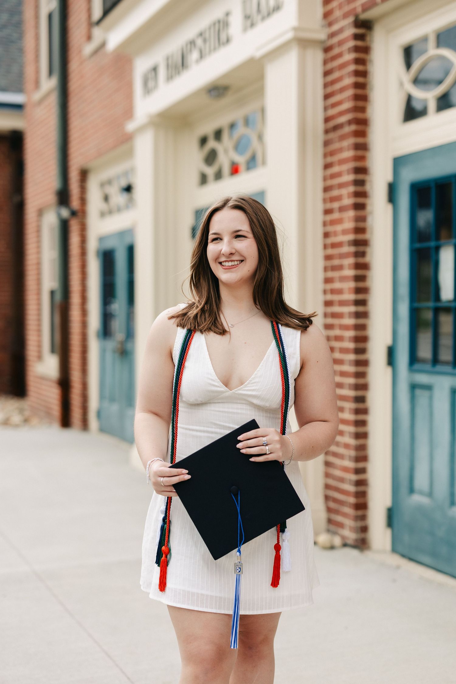 Graduate in white dress holds decorated cap while walking through brick-lined street.