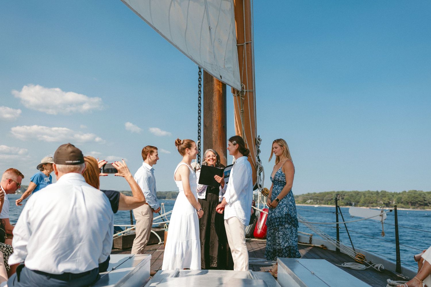 Wedding ceremony taking place on the deck of a sailboat with guests gathered around on a beautiful day.