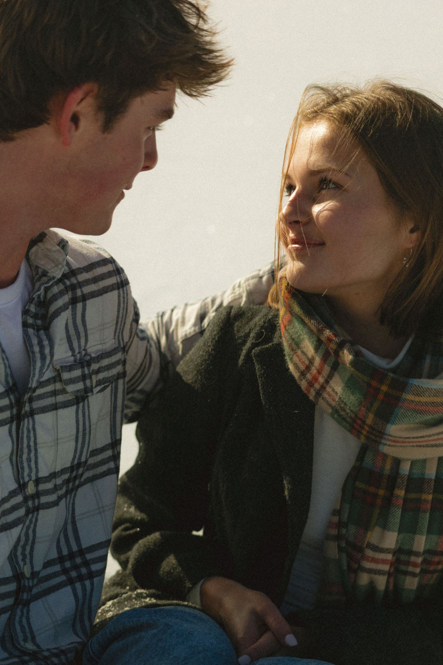 A young couple smiles at each other outdoors, wearing plaid shirts and a scarf on a bright day.