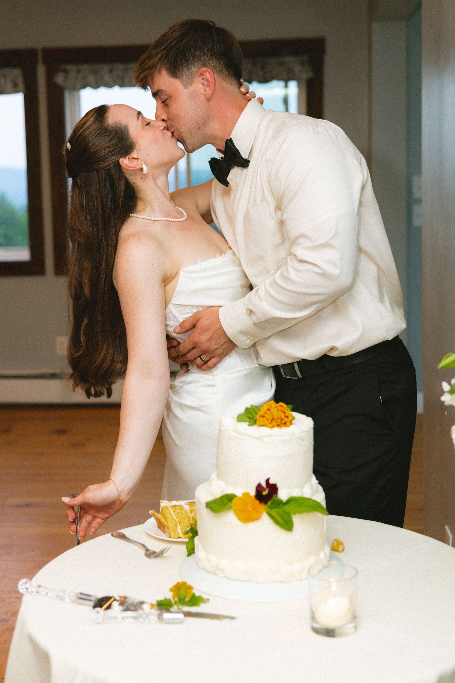 Bride and groom sharing a kiss while cutting their two-tier white wedding cake at reception.