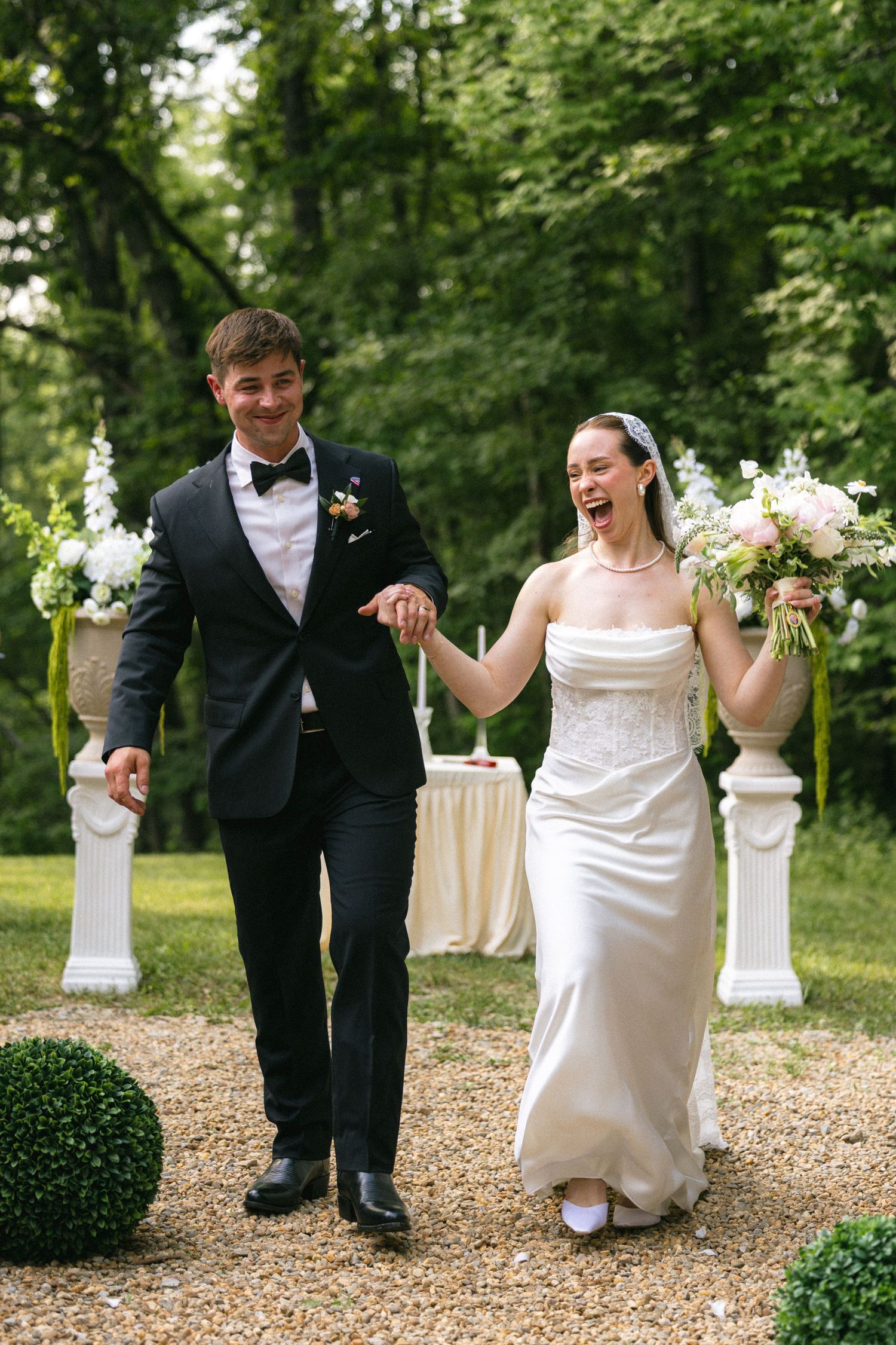 Joyful bride and groom walking hand in hand after outdoor wedding ceremony surrounded by lush green trees.