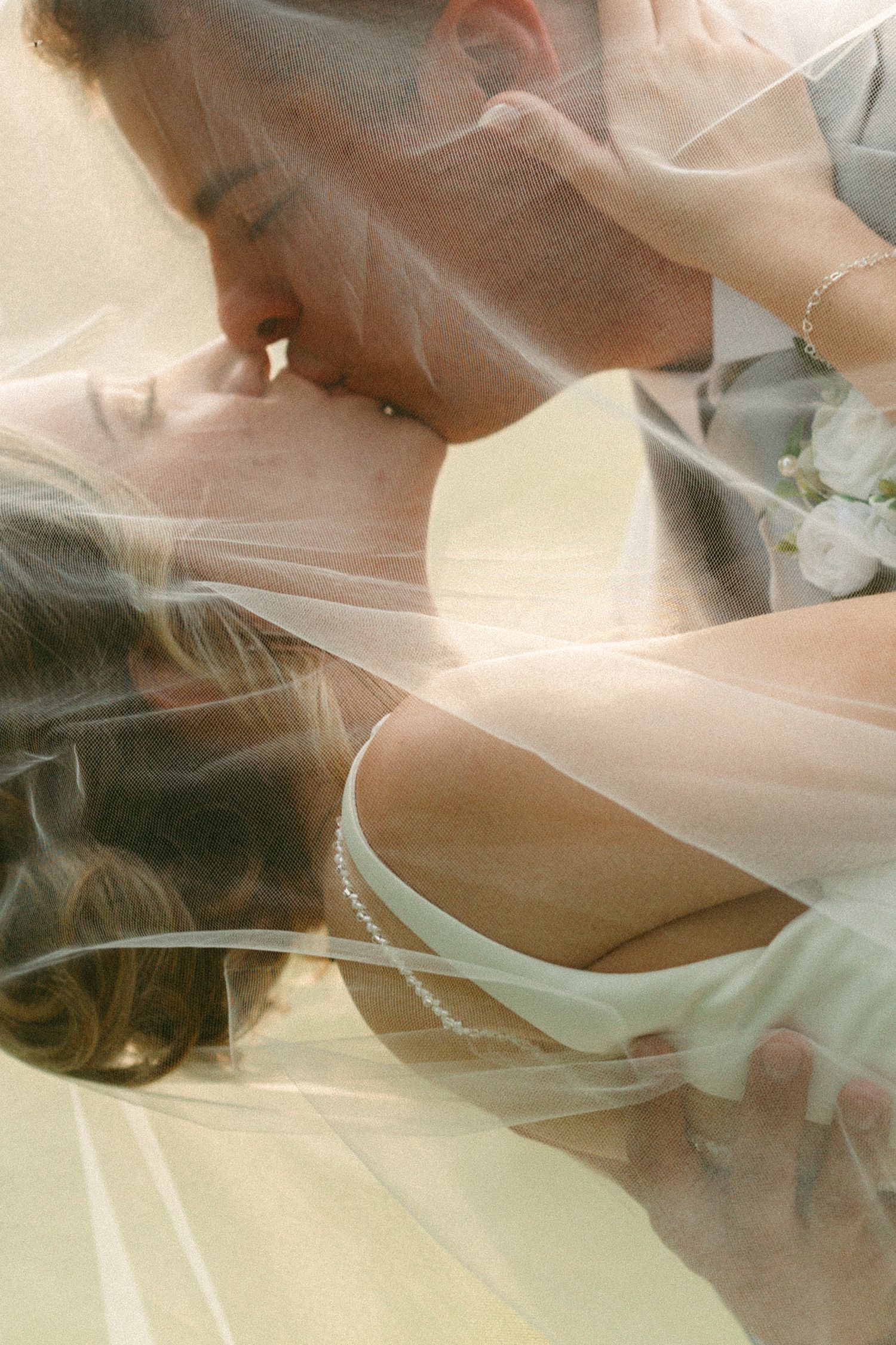 A bride and groom share a romantic kiss beneath a flowing wedding veil in a soft, dreamy photograph.