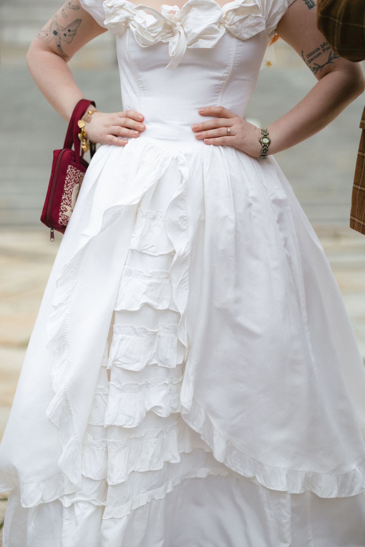 Woman in white ruffled ball gown wedding dress with bow detail, holding red velvet clutch purse, tattoos visible on arms.