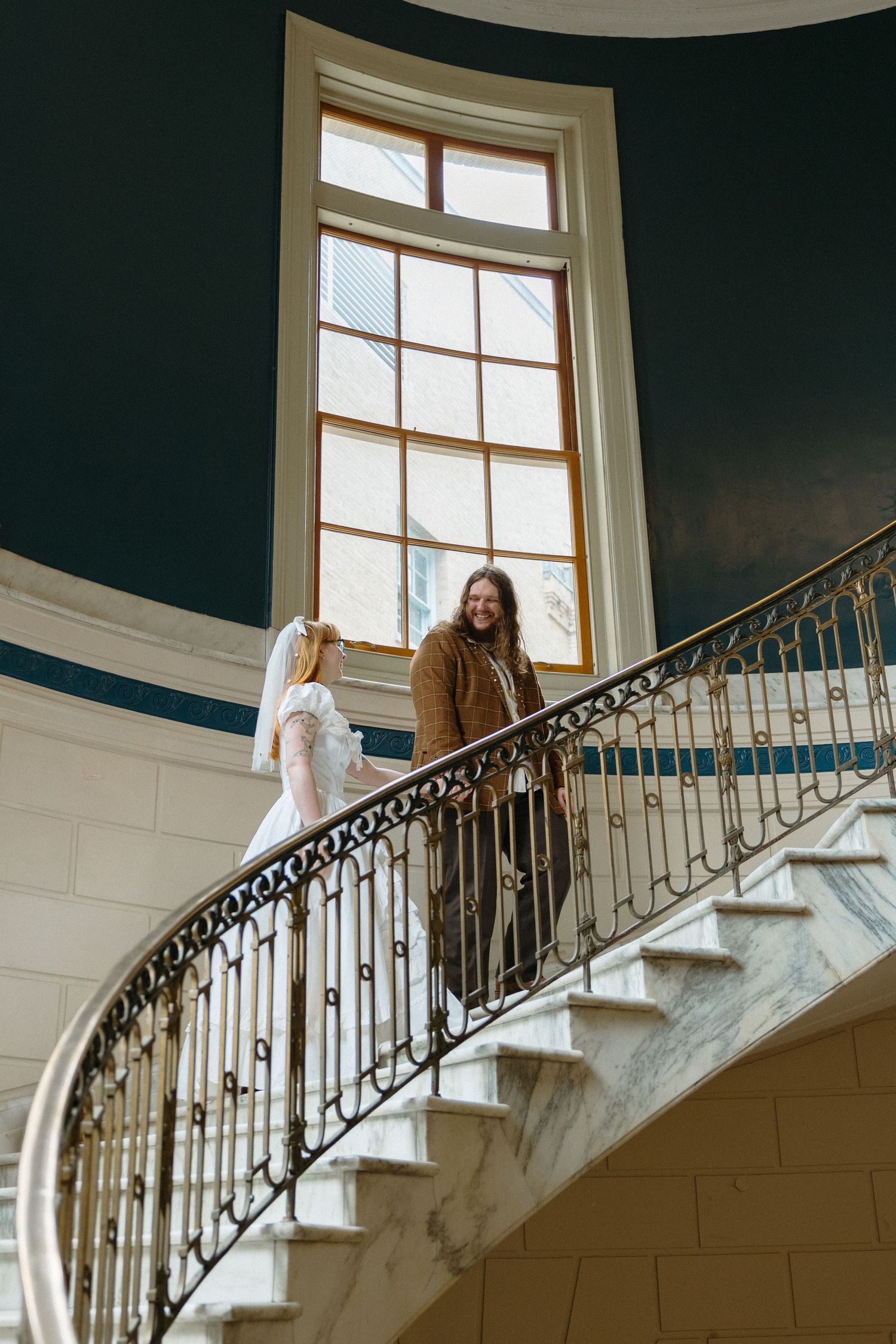 Bride and groom share a romantic moment on an elegant curved staircase with ornate gold railing in a grand venue.