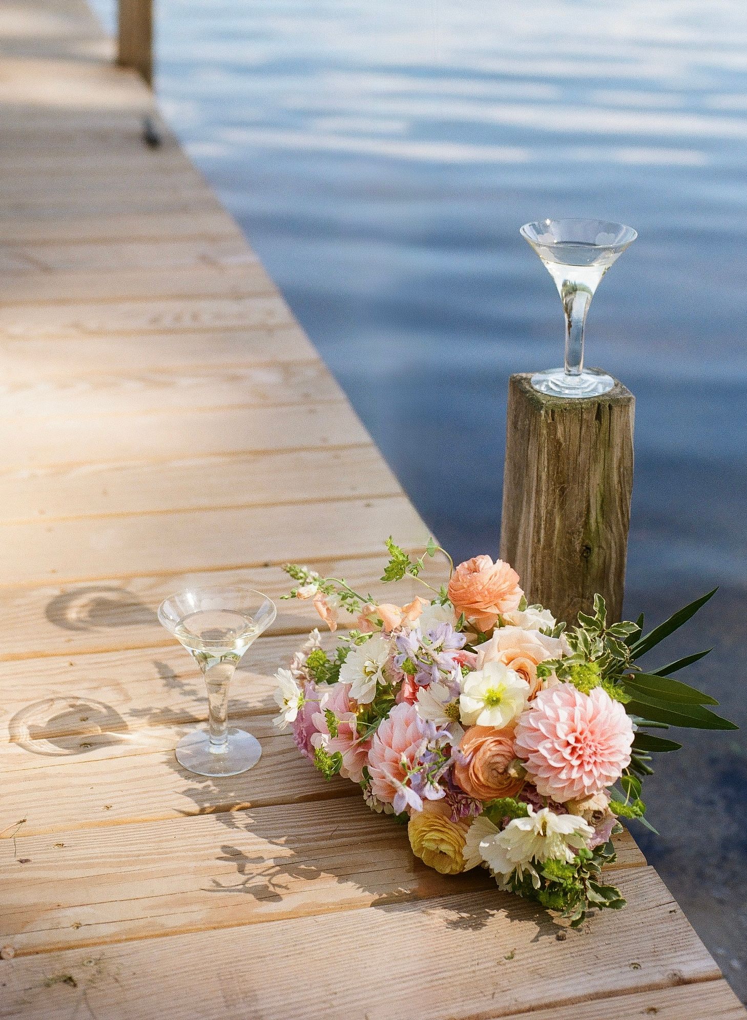Floral bouquet with pink dahlias and roses beside martini glasses on a wooden dock overlooking calm blue water.