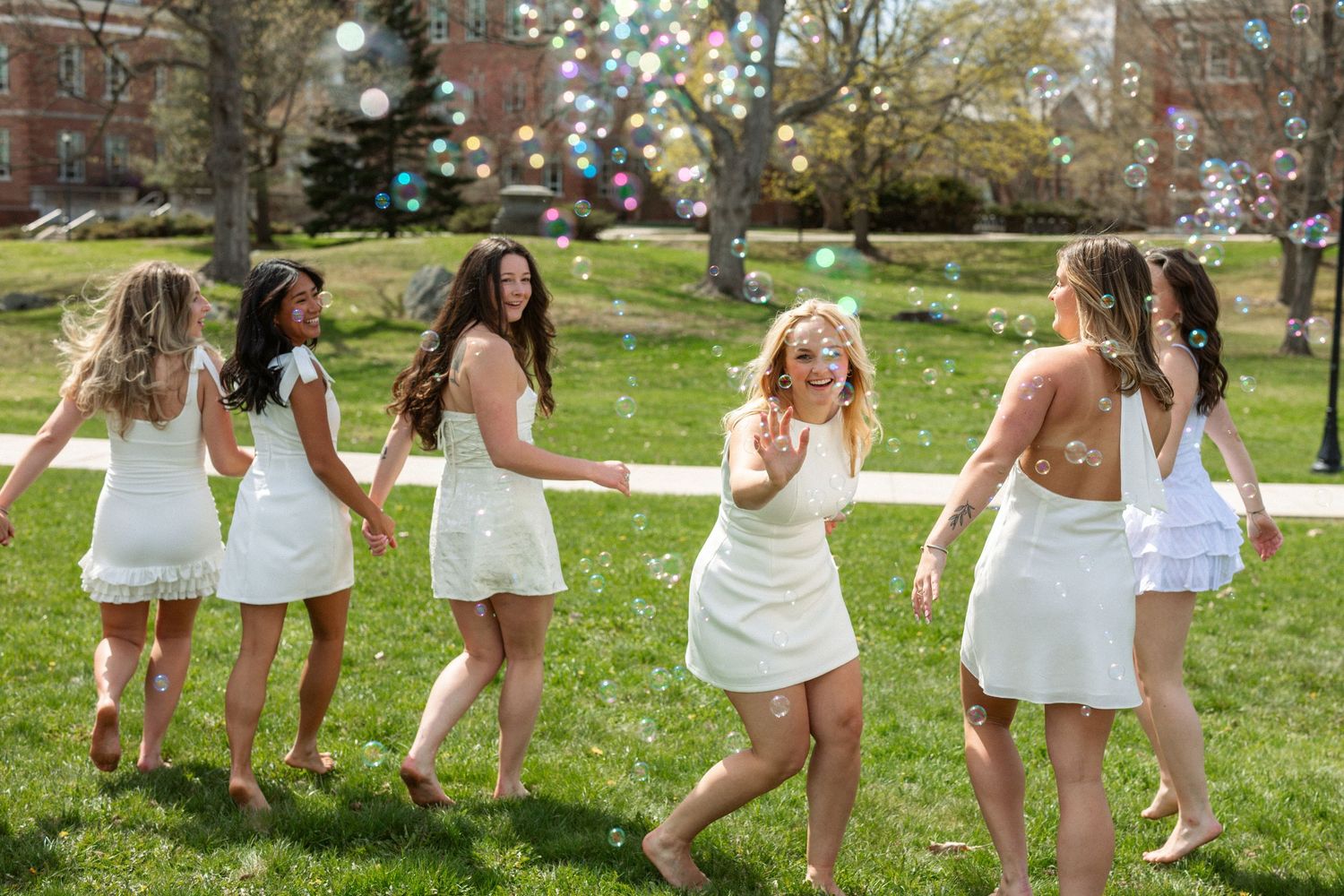 Group of young women in white dresses dancing on green grass with bubbles floating in the air on a sunny day.