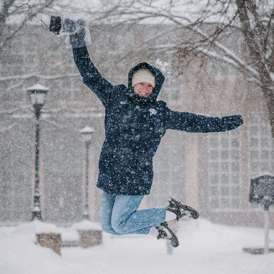 Person jumping joyfully during snowfall on a winter street with lampposts.