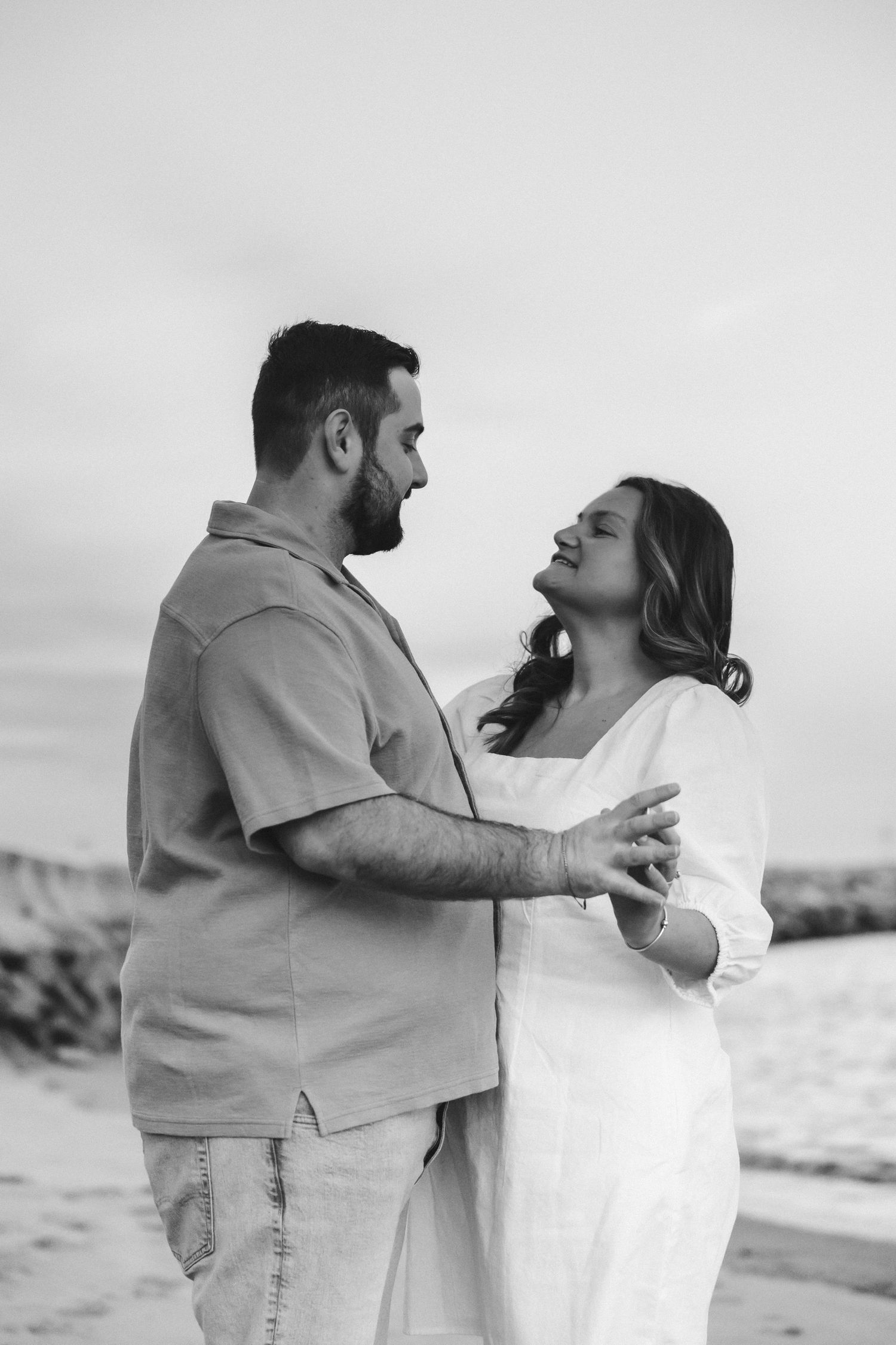 Black and white photo of a couple embracing on a beach, gazing at each other romantically at sunset.
