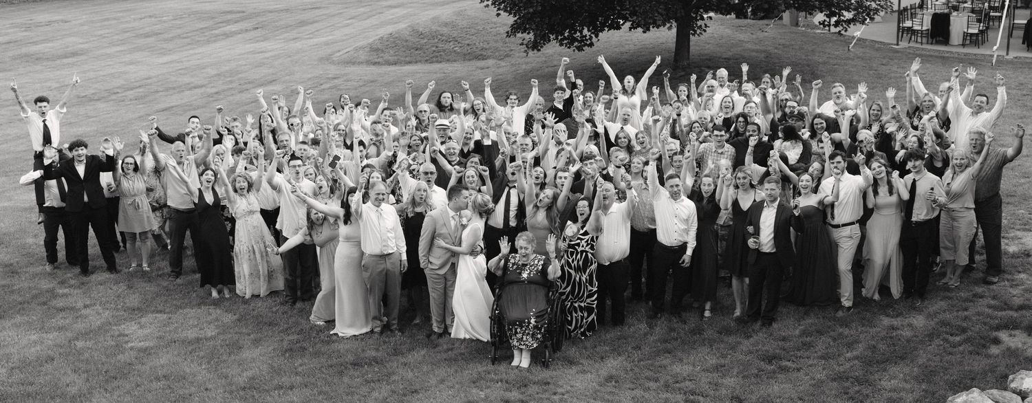 Large wedding group photo outdoors with guests raising arms in celebration, black and white panoramic shot.
