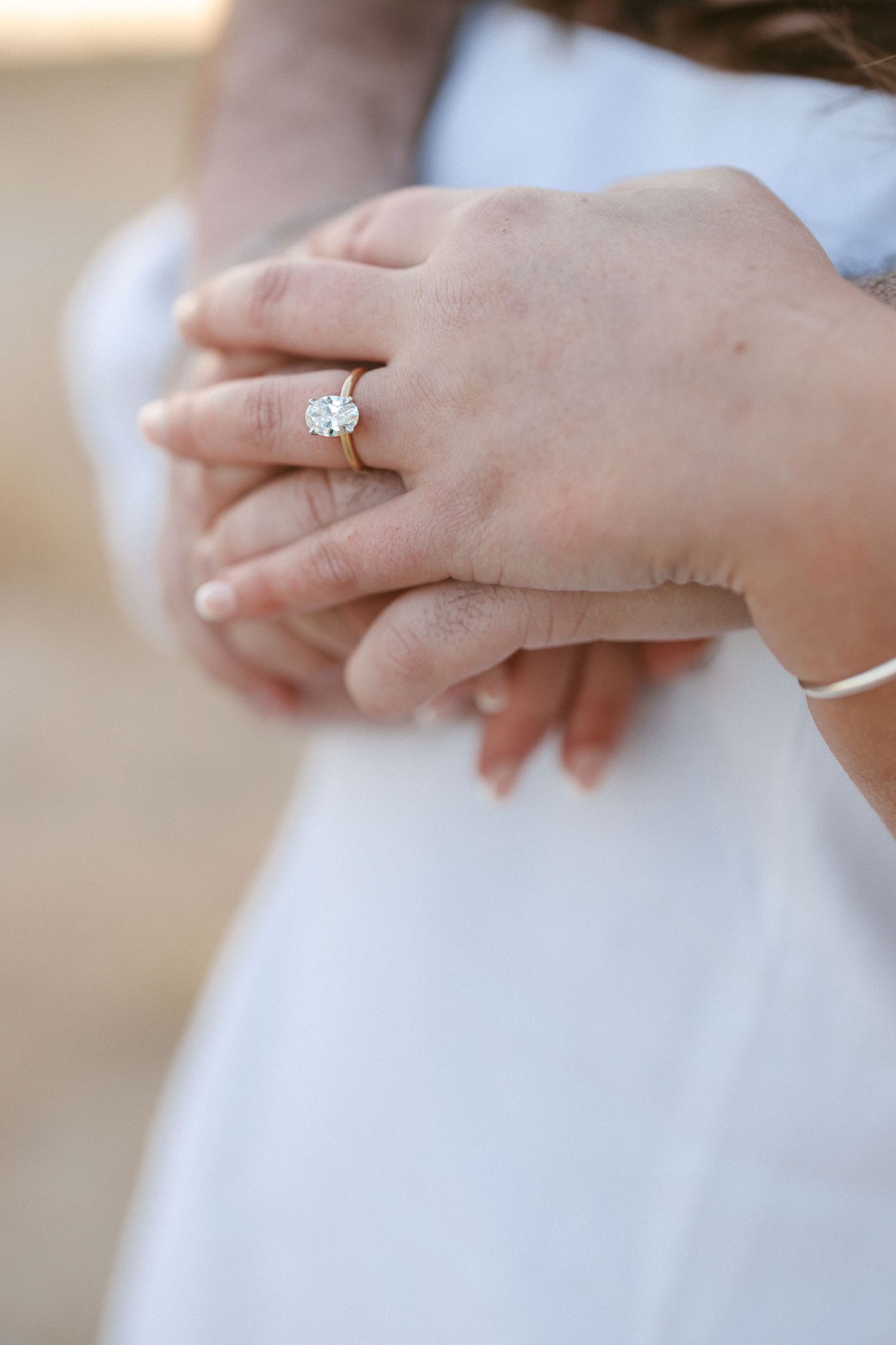 Close-up of hands clasped together showing a sparkling diamond engagement ring on a gold band against a white dress.