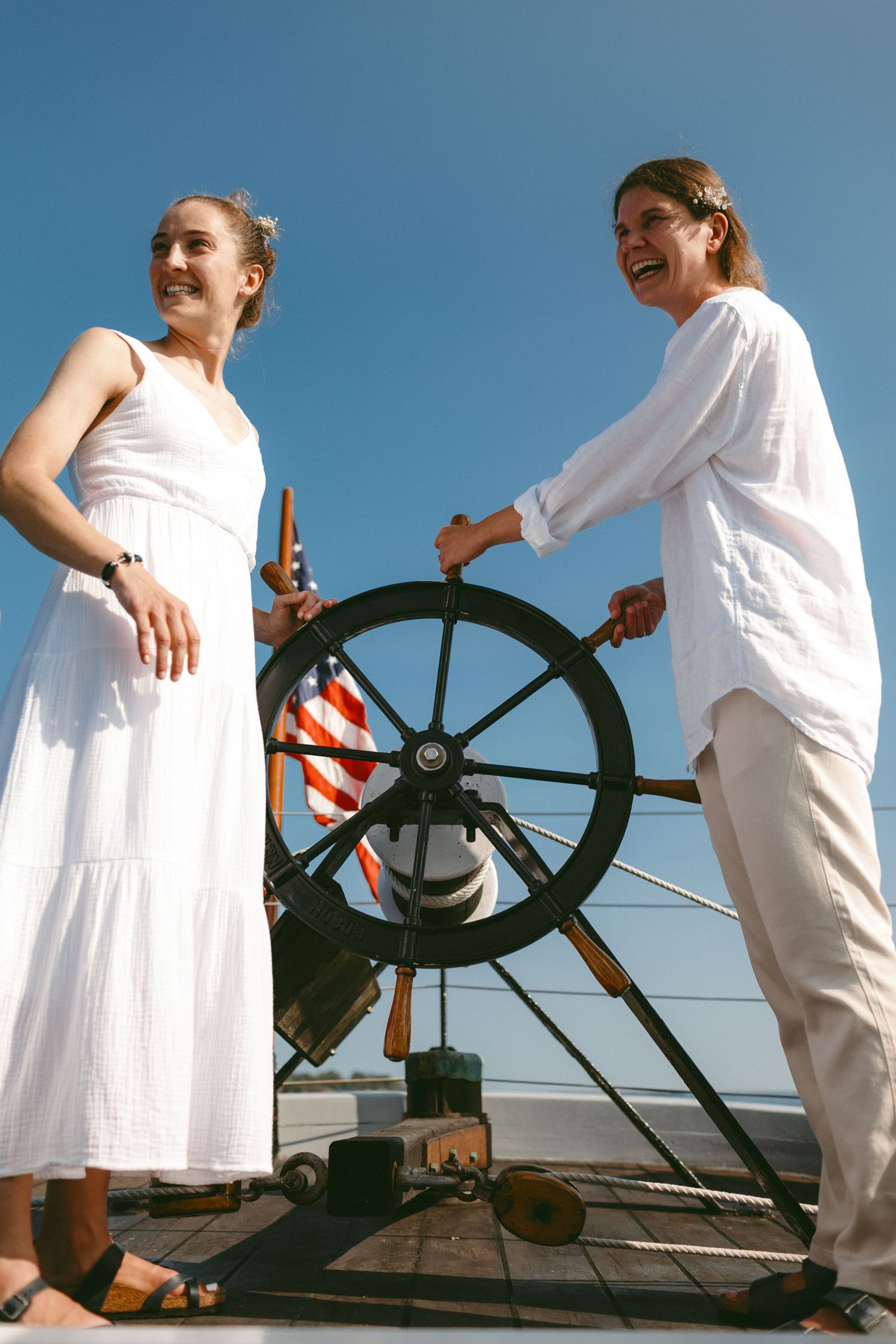 Two people in white outfits standing at a ship's wheel against blue sky.