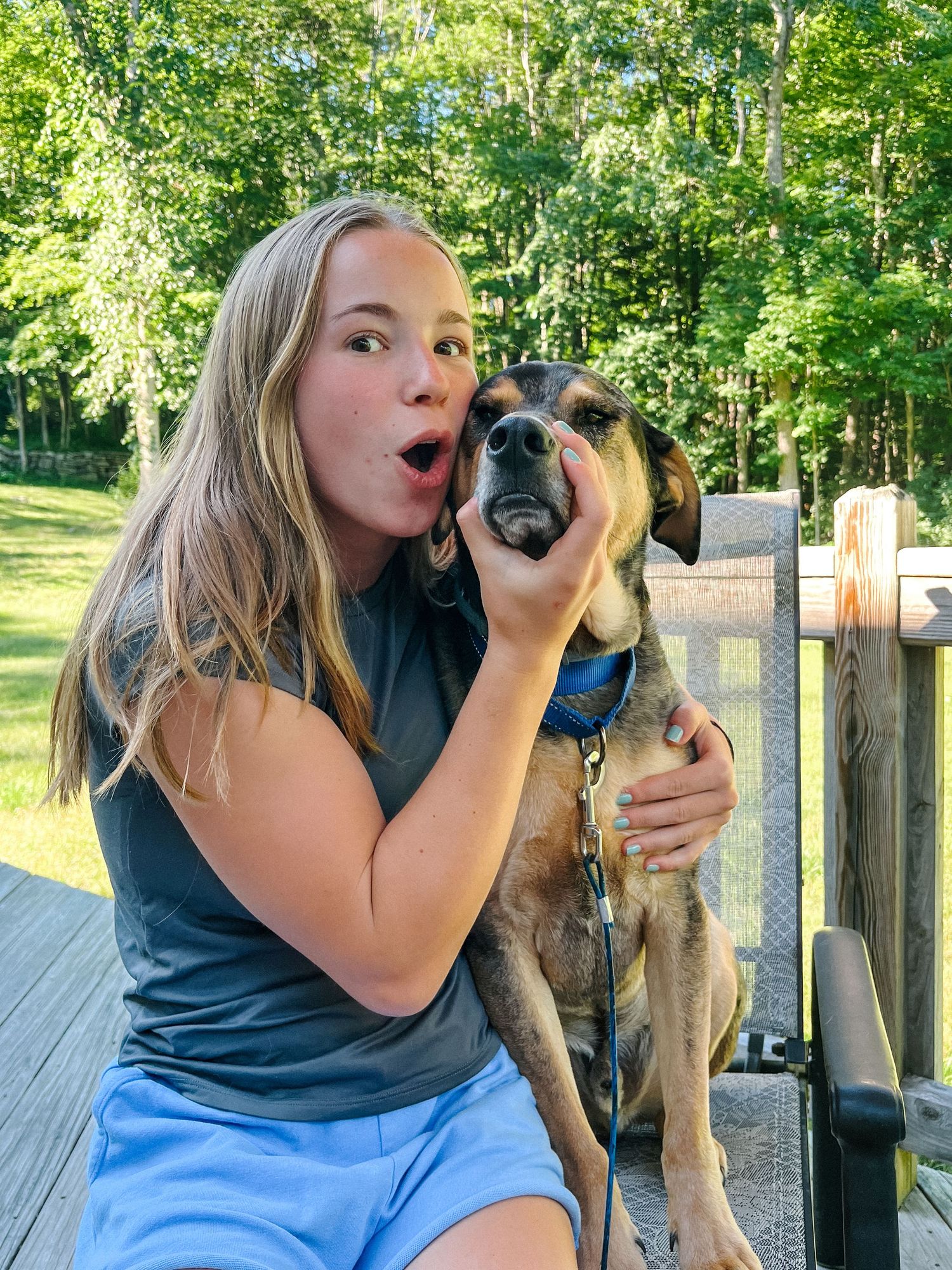A playful moment on a wooden deck shows someone making a silly expression while posing with their mixed-breed dog outdoors.