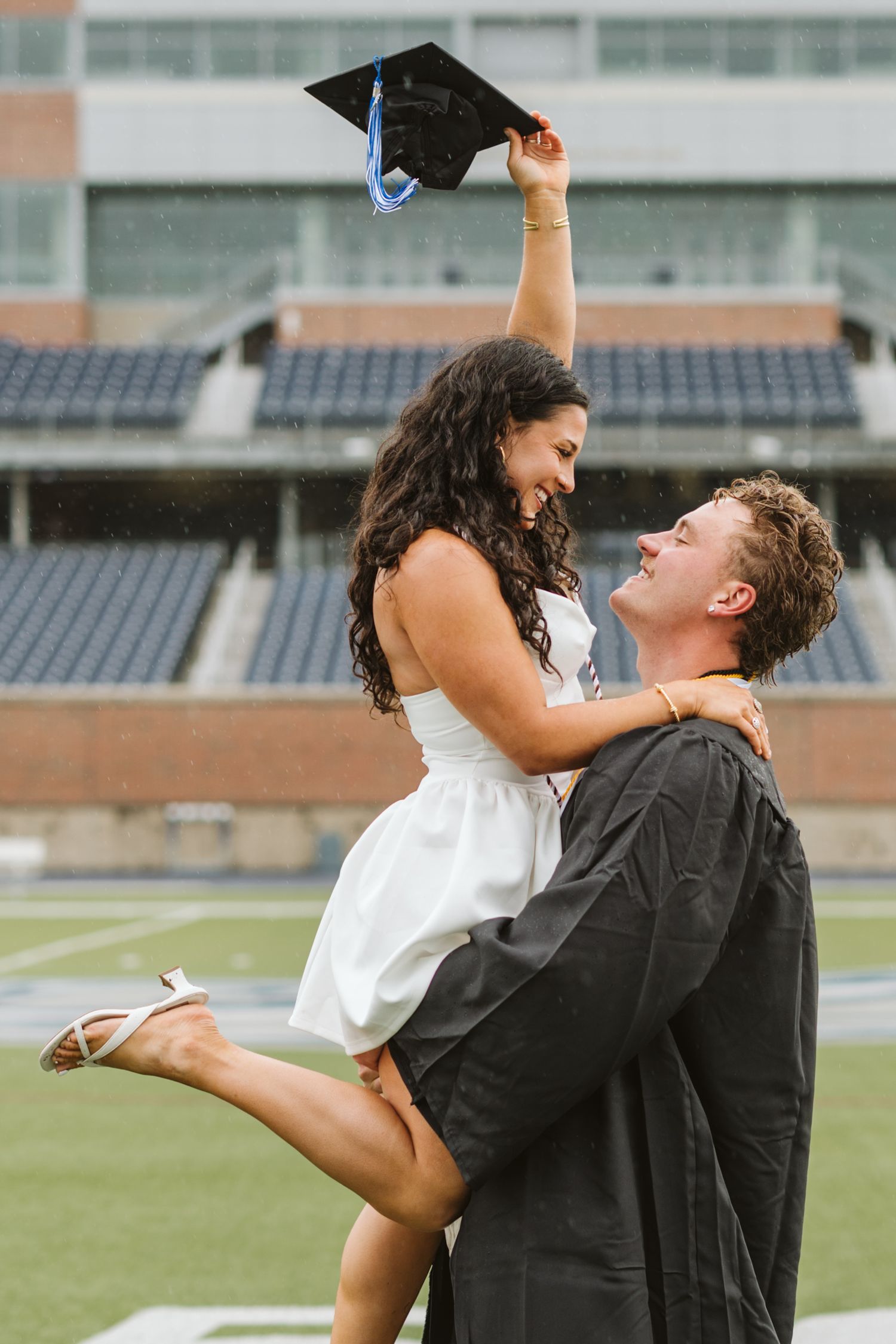 A man in graduation gown lifts a woman in white dress holding a cap at a stadium.