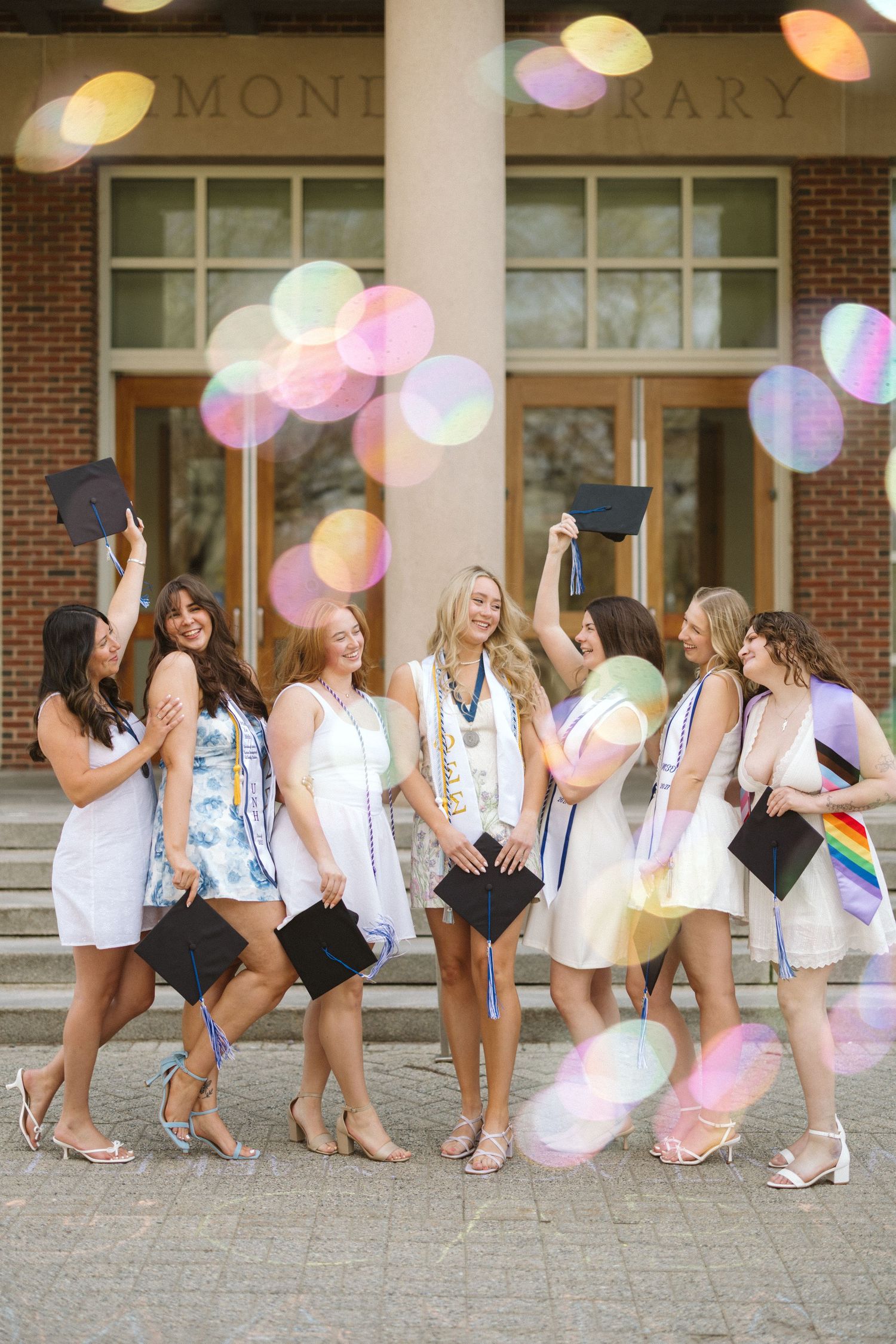 Seven college graduates in white dresses celebrate outdoors, tossing caps with colorful bokeh bubbles in front of a brick library.