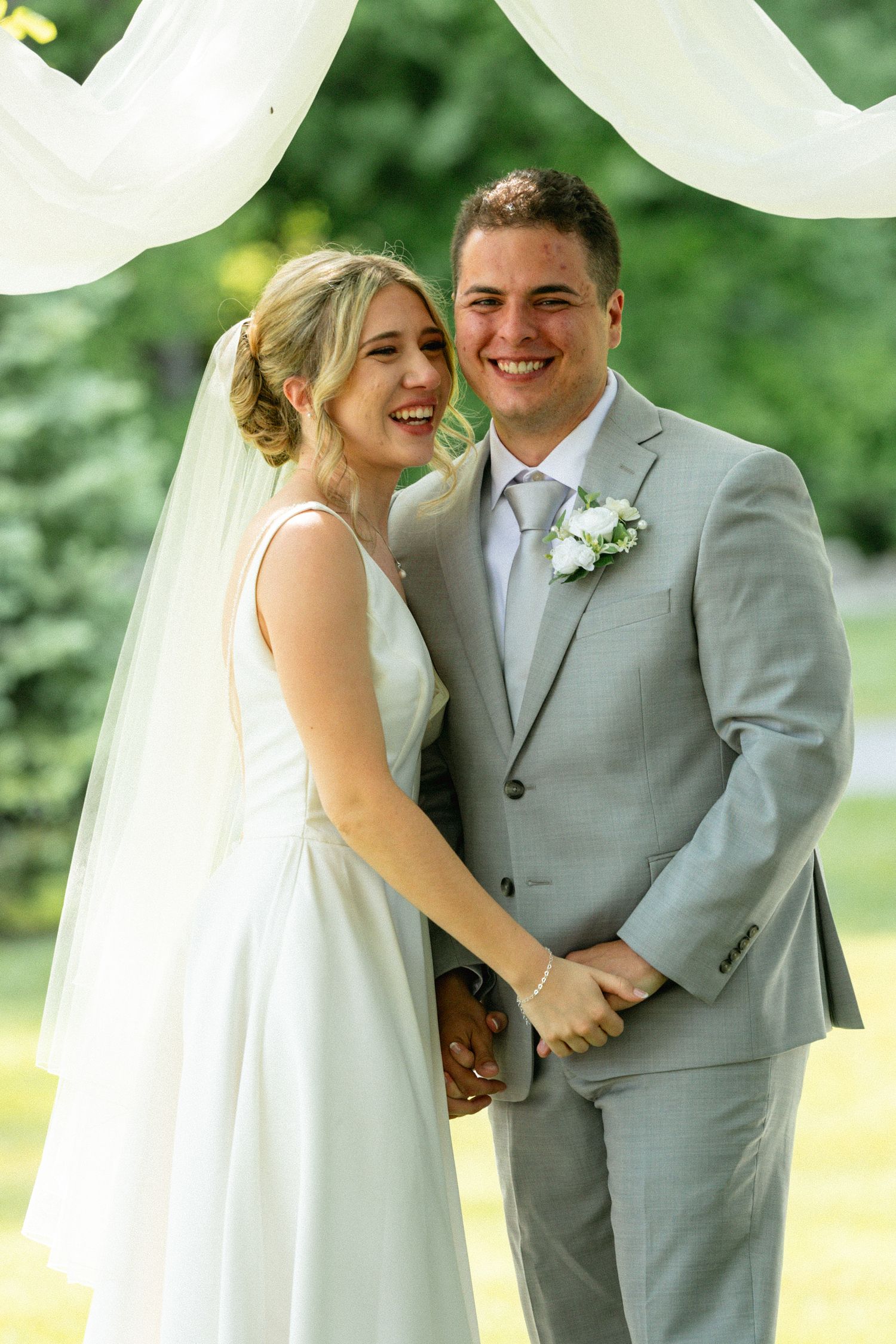Smiling bride and groom pose together outdoors under white draped arch on their wedding day.