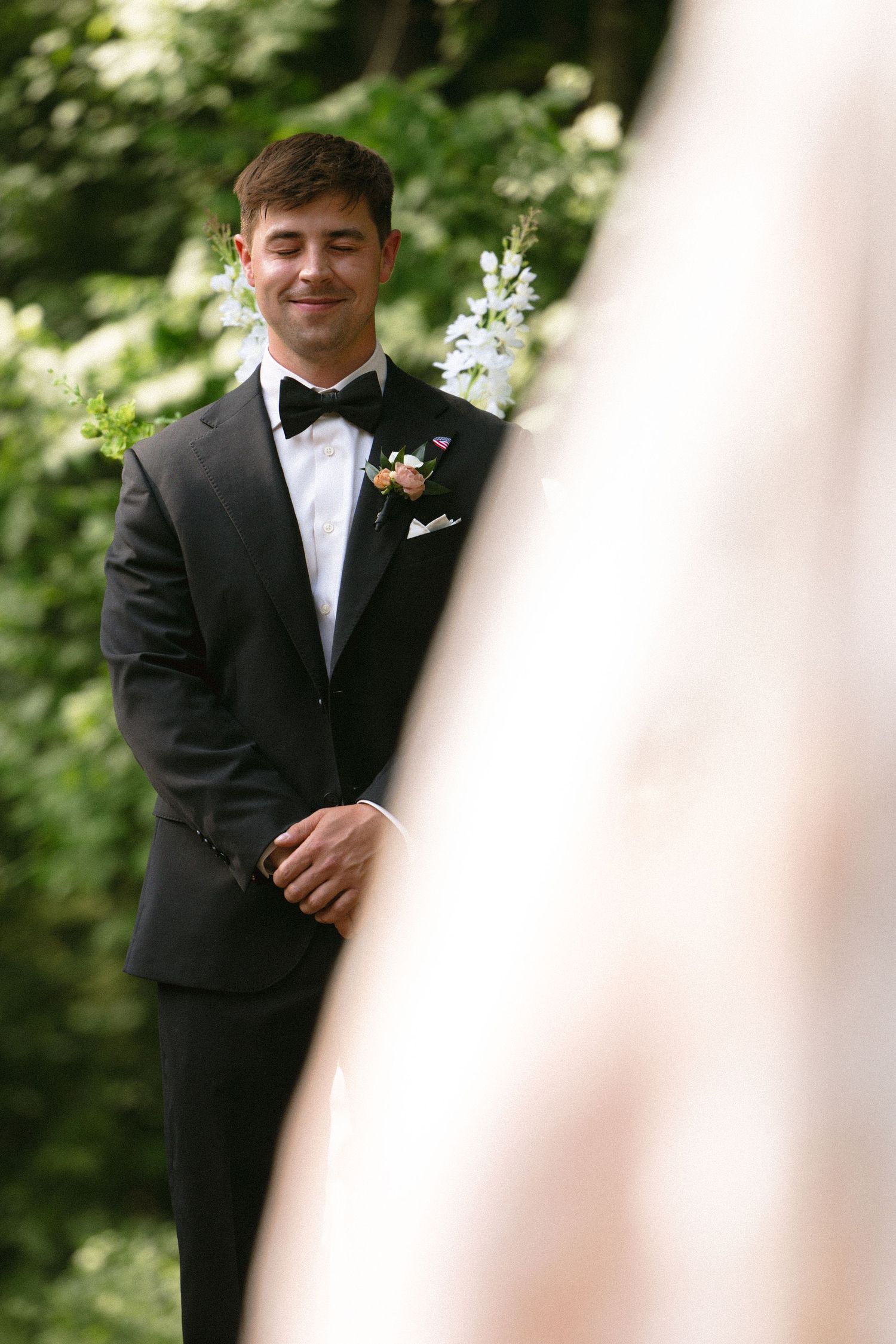 Smiling groom in black tuxedo with boutonniere watching bride approach at outdoor ceremony.