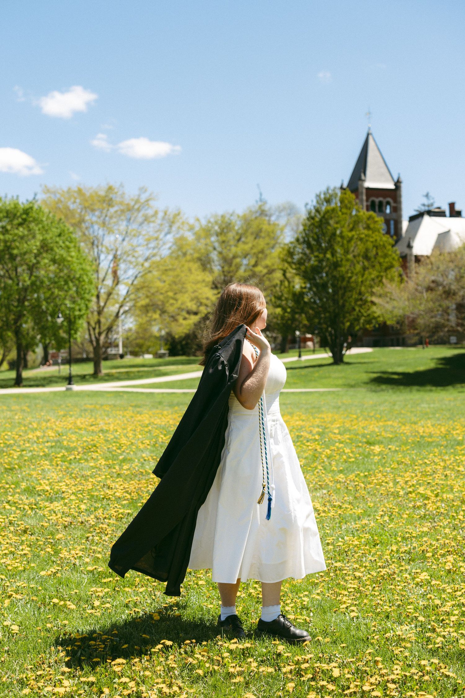 Young woman in white dress holding graduation gown on a sunny green campus lawn with yellow wildflowers.