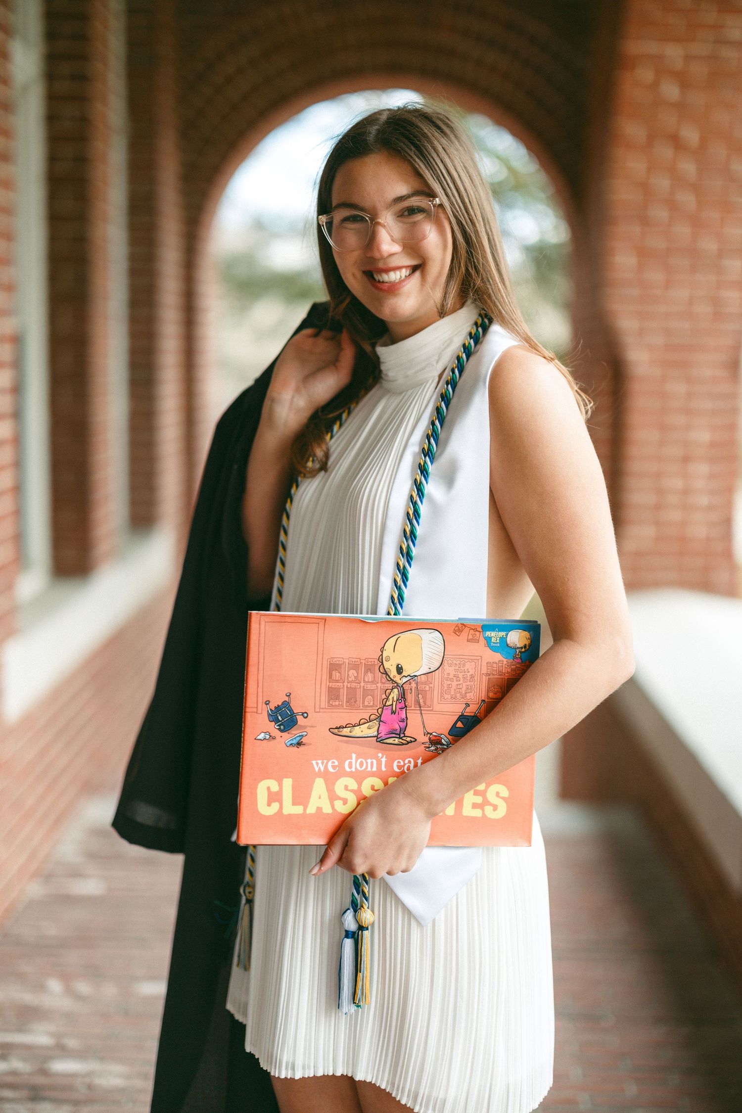Smiling graduate in white dress holds Peanuts book and tassels with black gown over shoulder in brick archway.