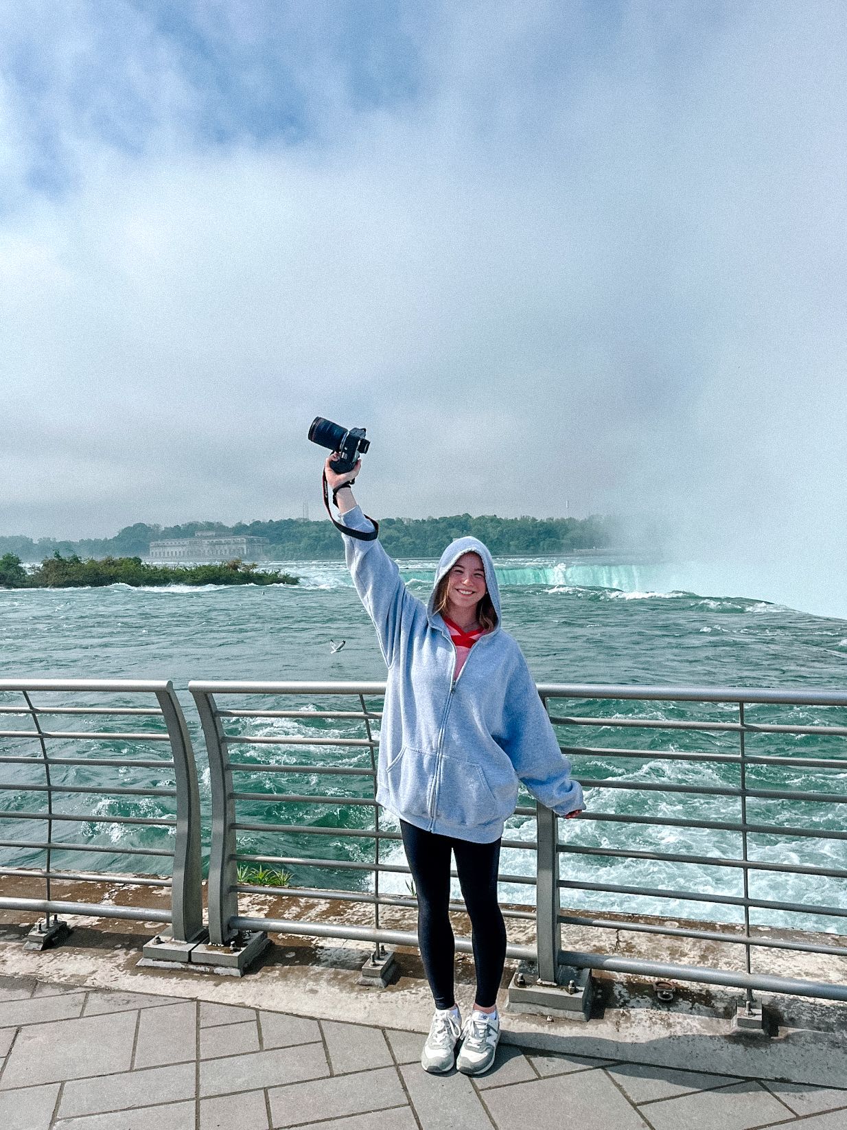Person in white jacket poses against dramatic backdrop of Niagara Falls on misty observation deck.