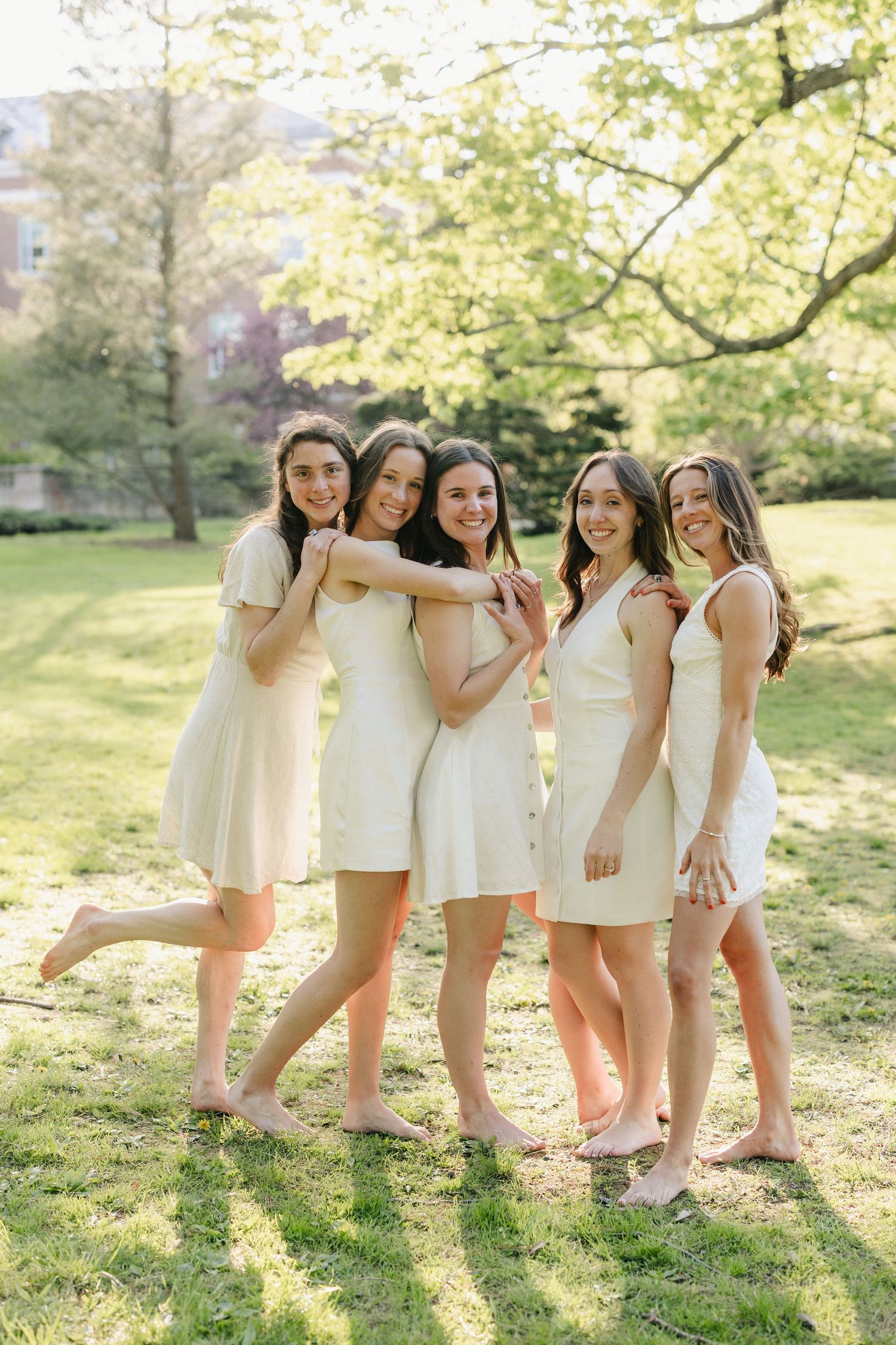 Group wearing white dresses poses playfully in sunlit garden setting.
