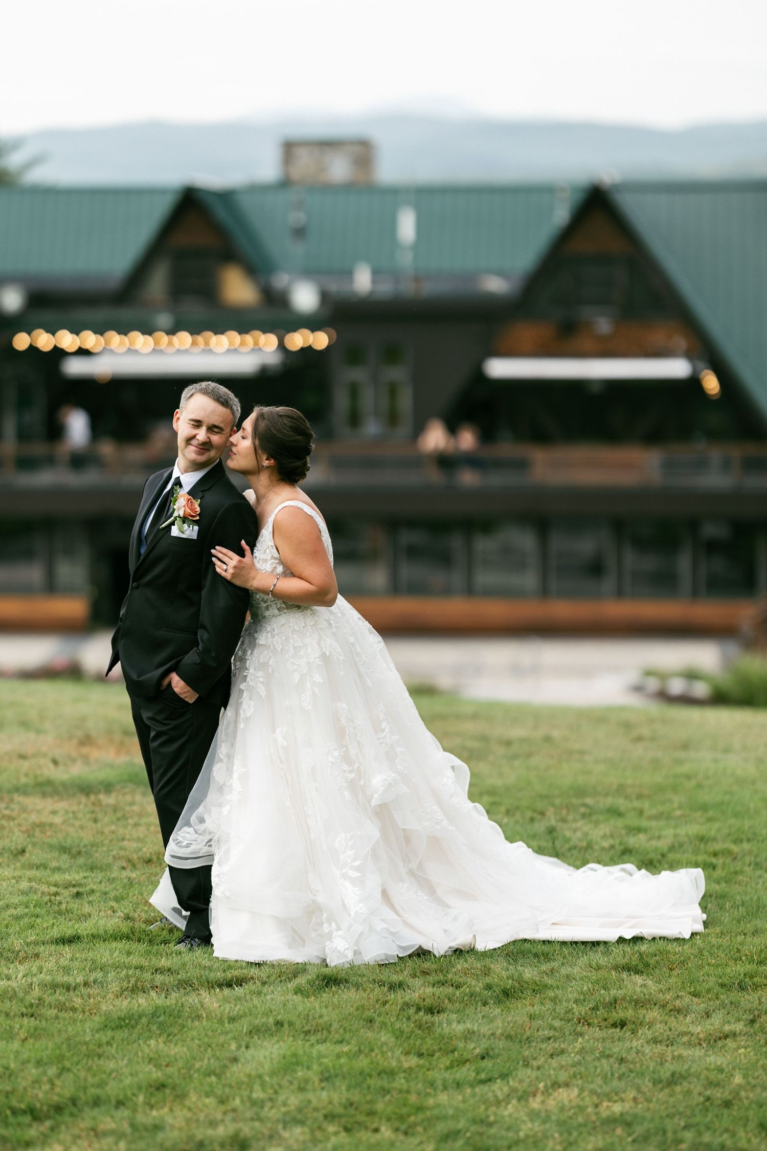A bride and groom share an intimate moment while sitting together on a grassy field at their outdoor wedding venue.
