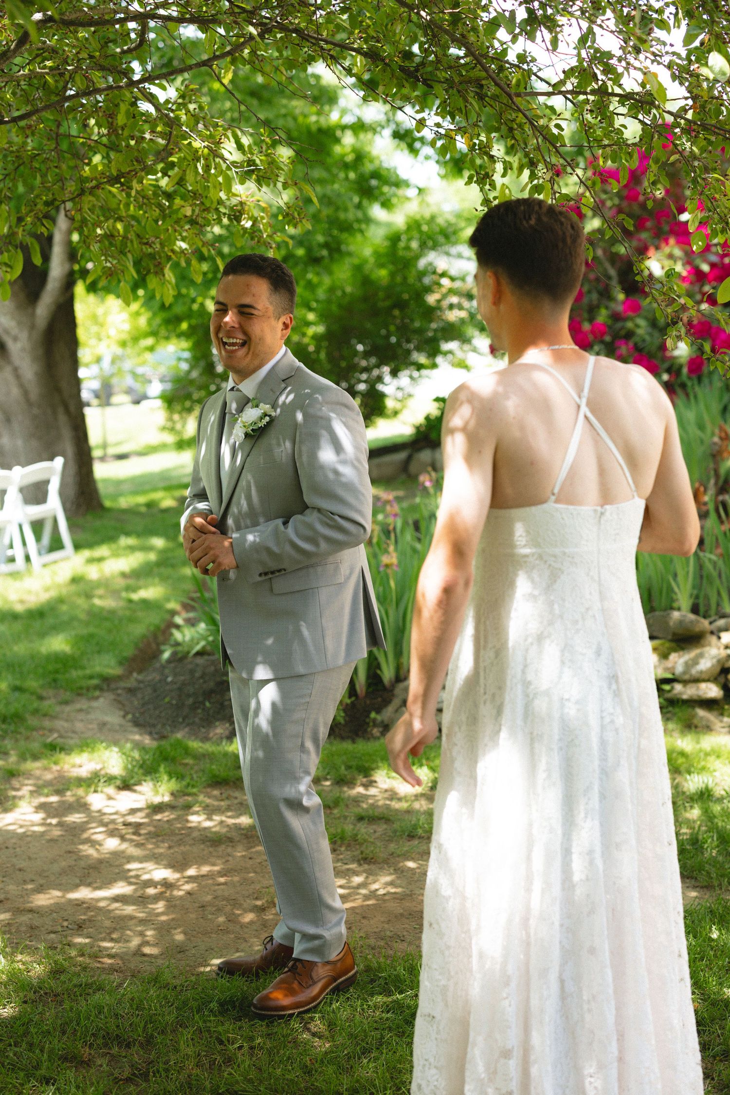 Groom in gray suit laughing during first look with bride in white dress in a sunny garden.