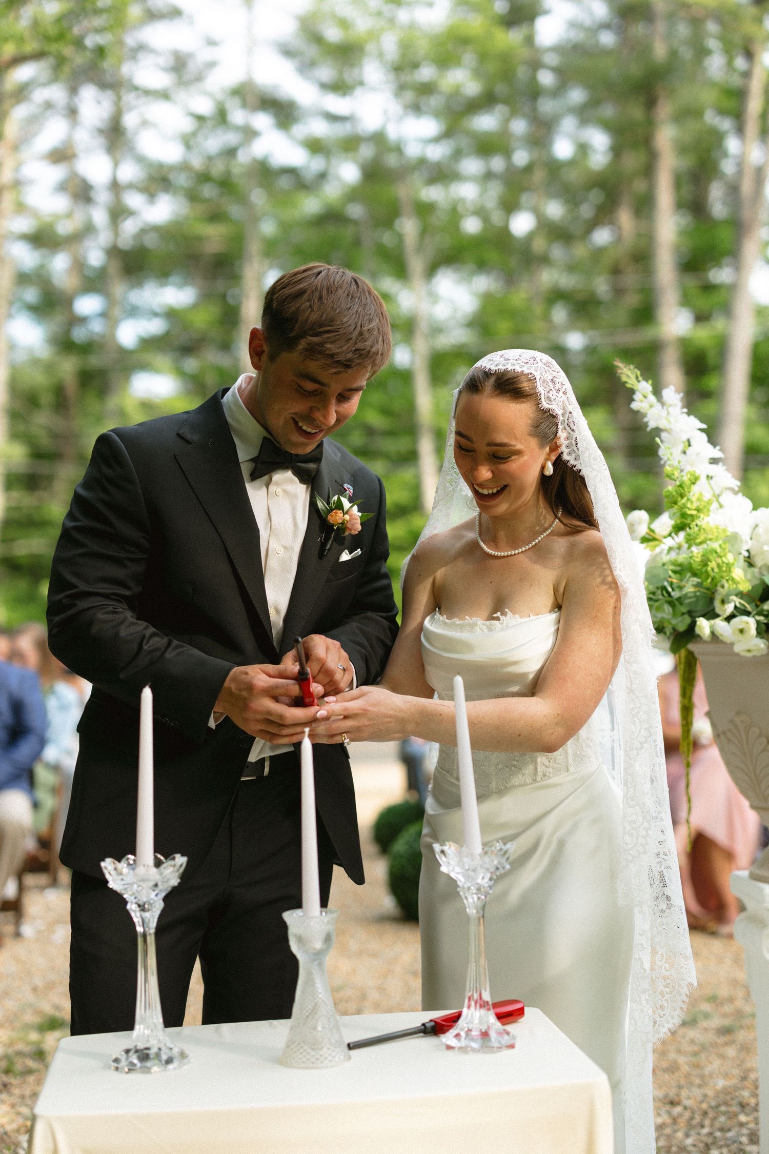 Smiling bride and groom perform unity candle ceremony outdoors surrounded by pine trees at their wedding.