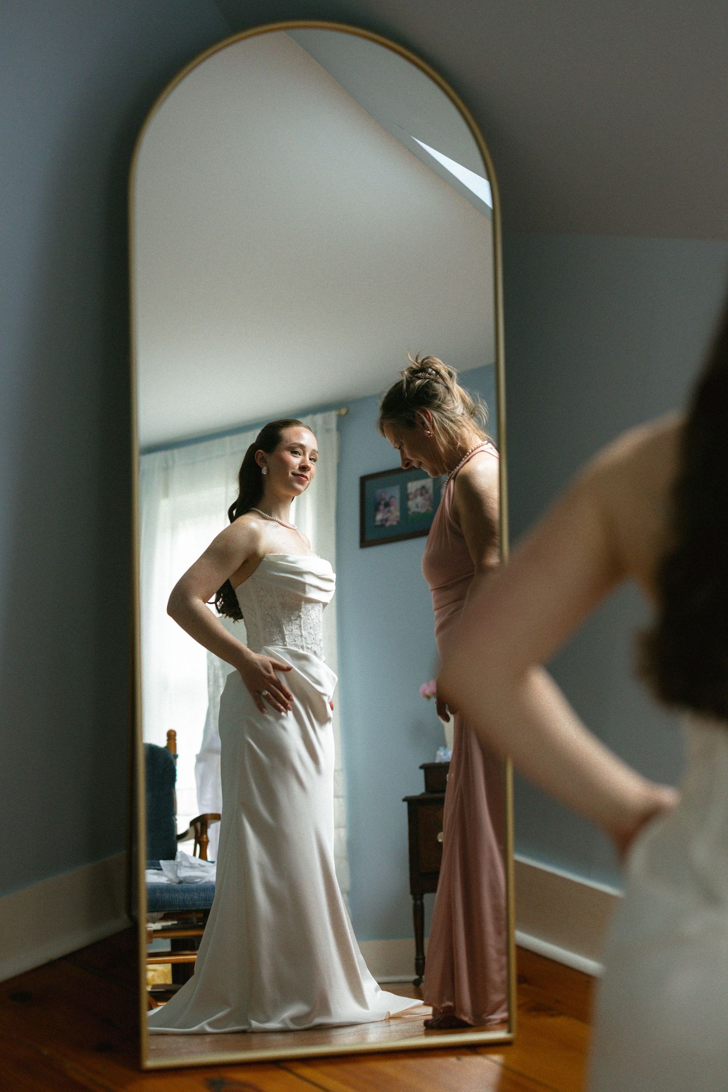 Bride in white strapless gown smiling at reflection in gold arched mirror while bridesmaid assists in blue room.