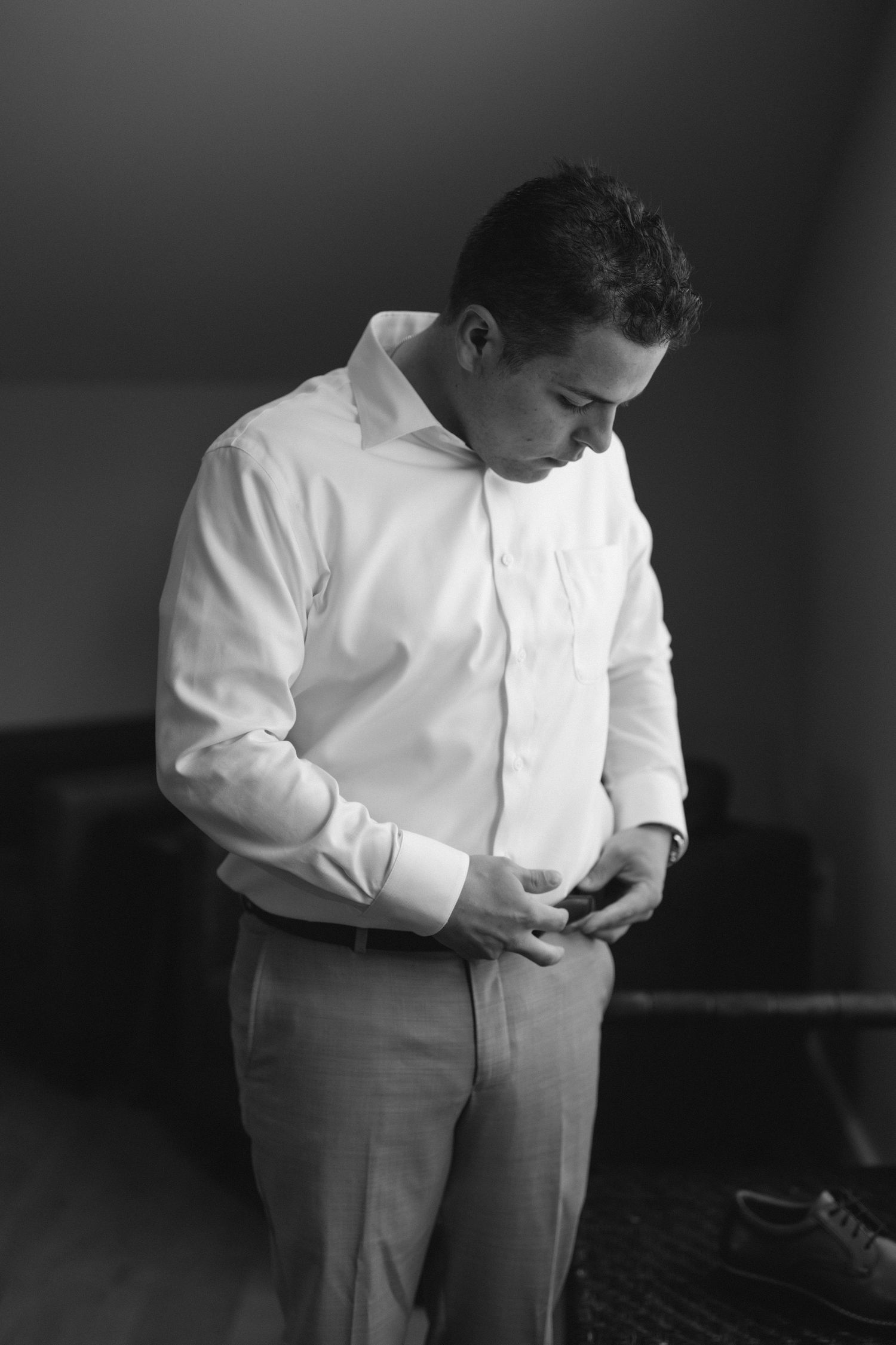 Black and white photo of a groom in a white dress shirt adjusting his belt while getting ready for his wedding.