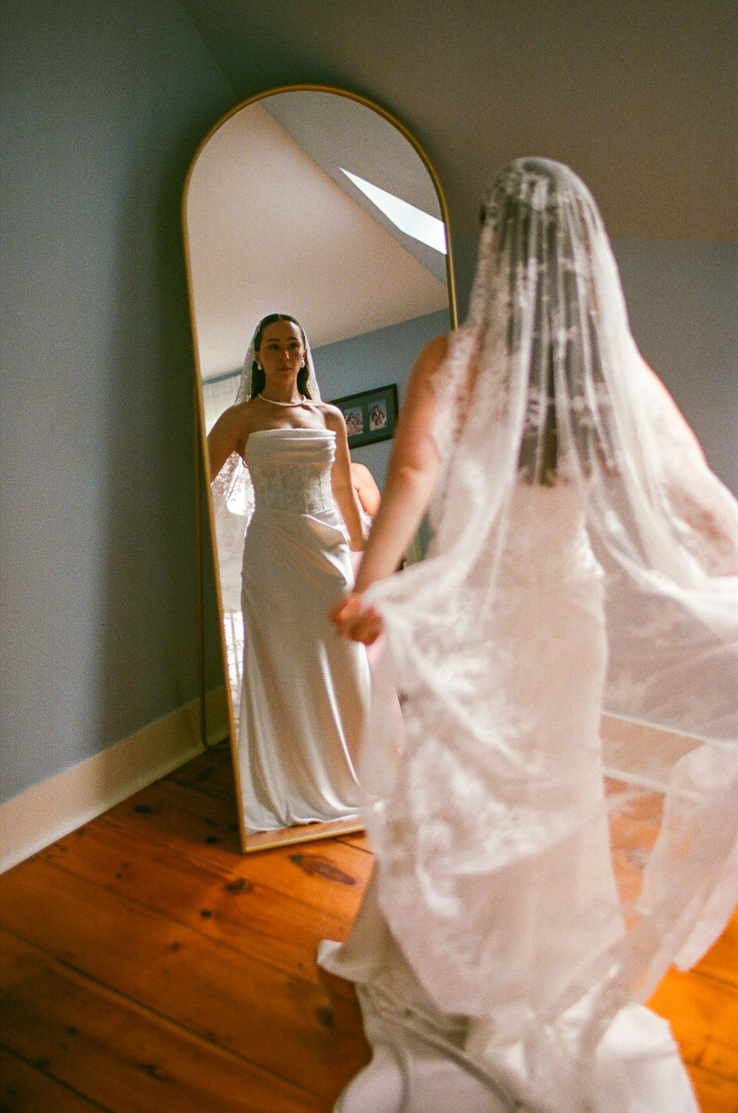 Bride in white gown and lace veil admires her reflection in a gold arched mirror on hardwood floors.