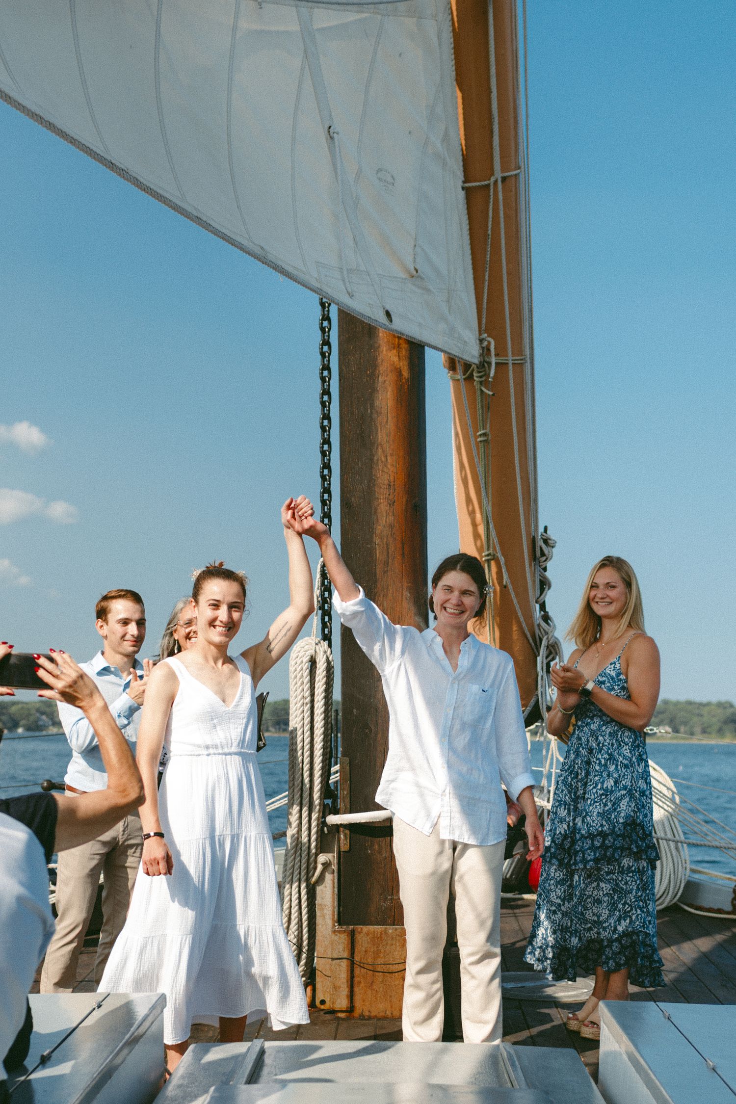 Celebratory moment on a sailboat deck with raised hands against white sails and blue sky.