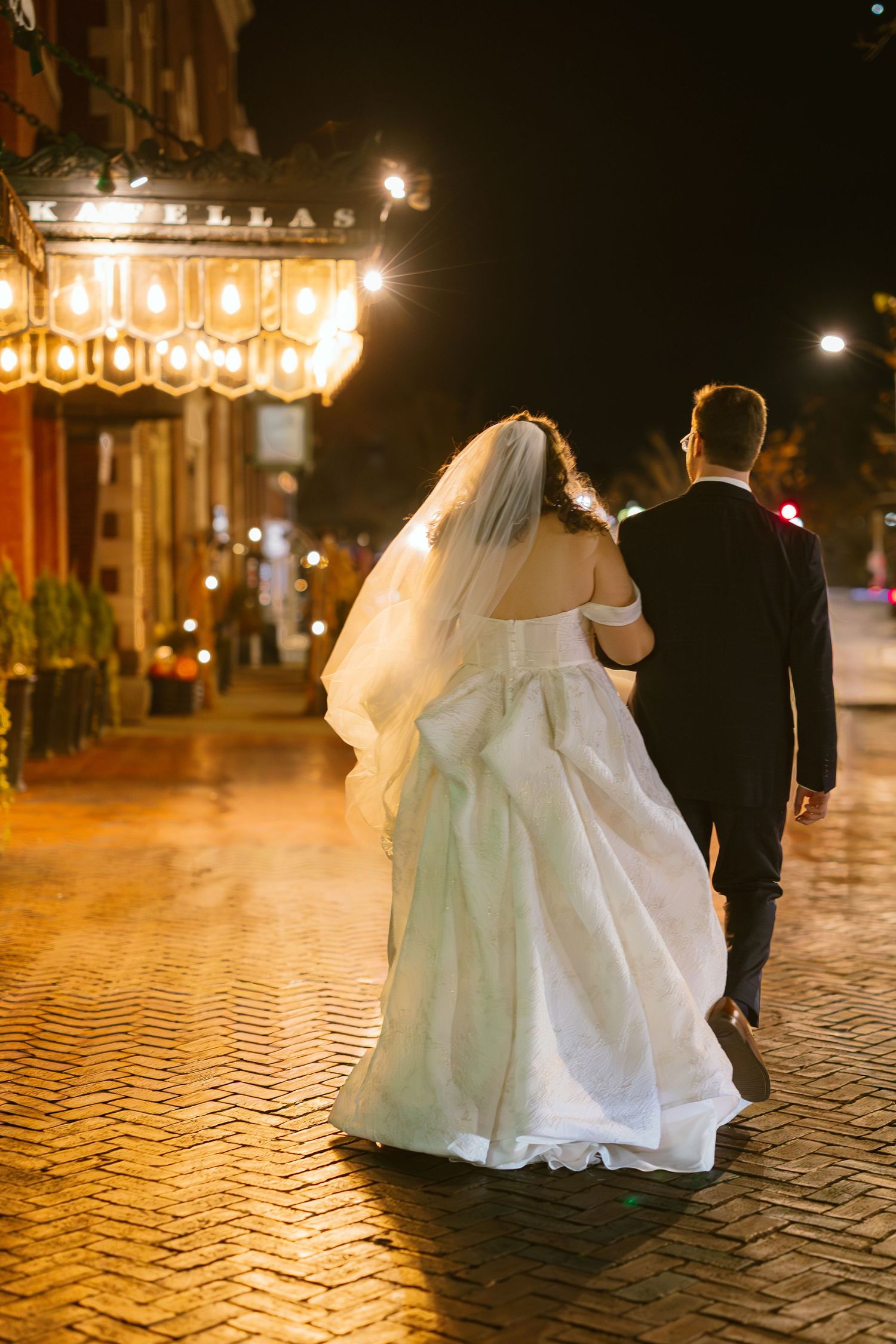 Newlyweds walk along a cobblestone street at night with glowing lights in the background.