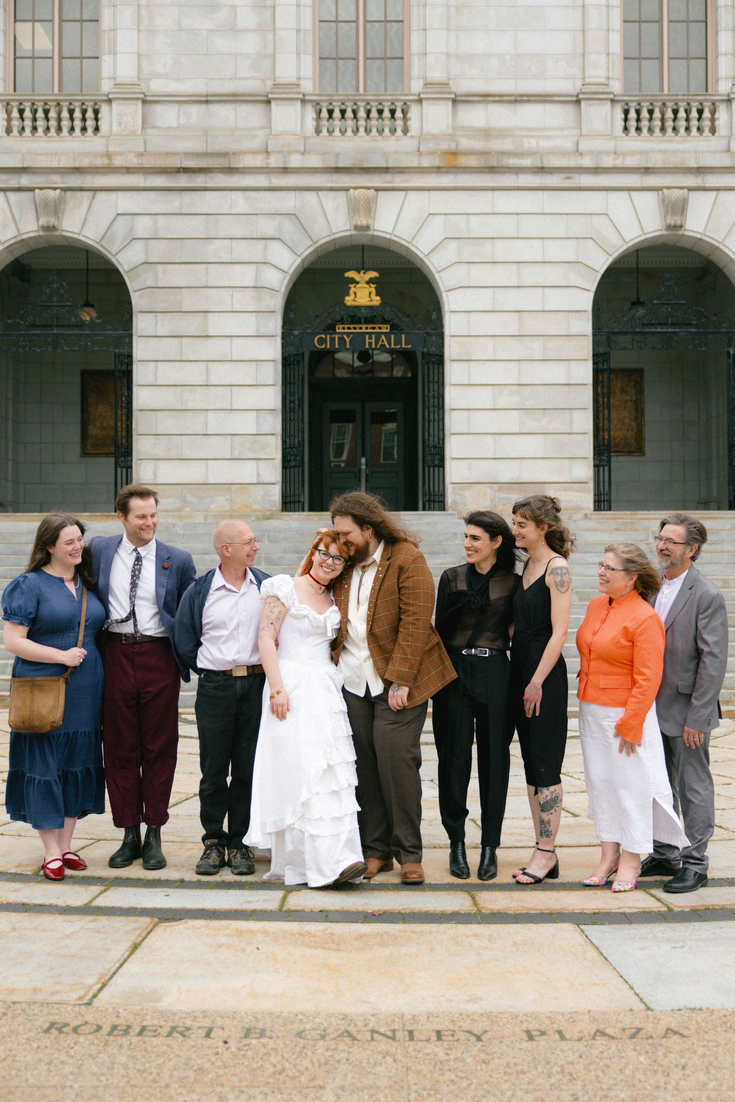 Wedding party poses joyfully outside Brooklyn City Hall on Robert F. Ganley Plaza on a sunny day.