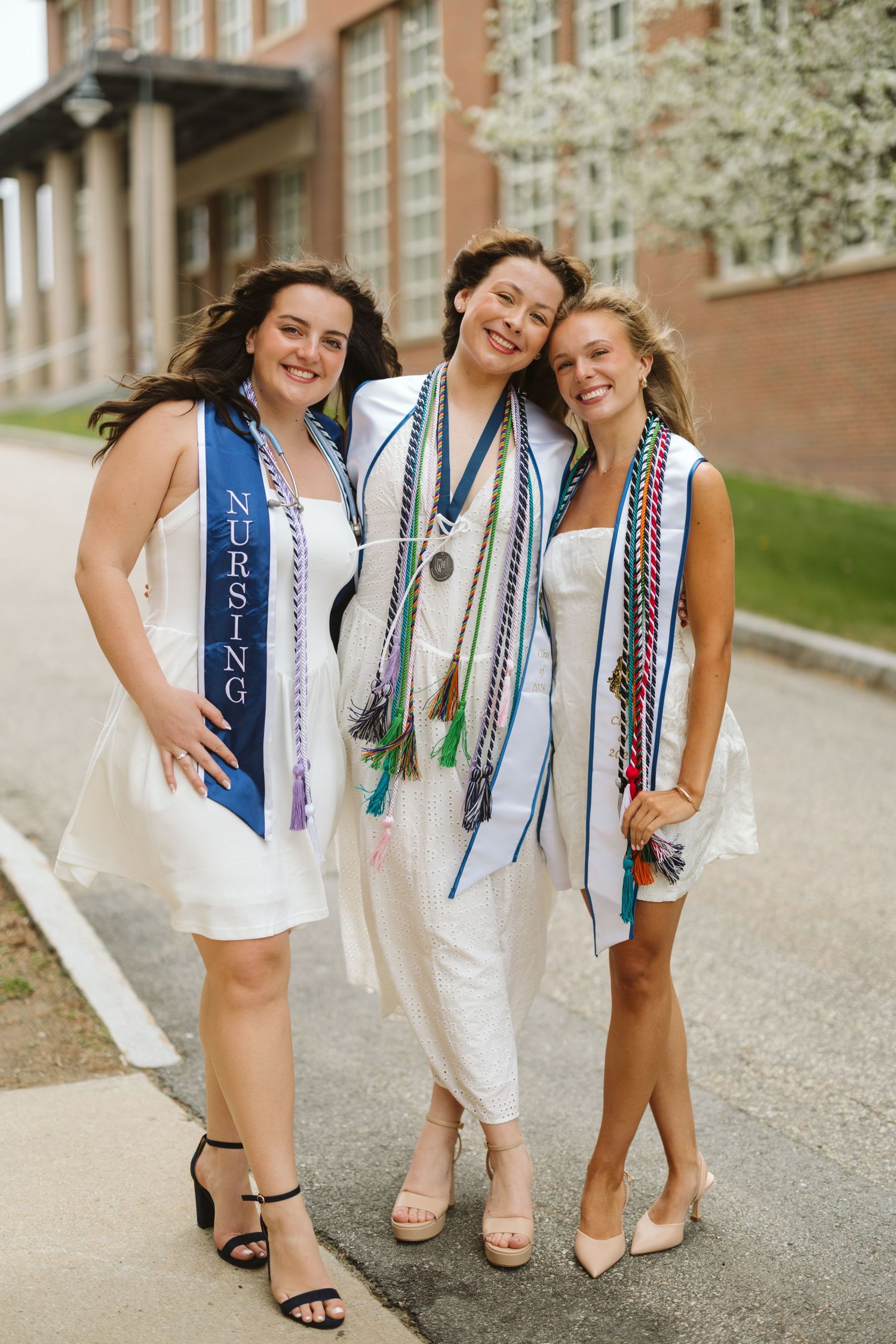 Three nursing graduates in white dresses with honor cords and sashes pose together outdoors on graduation day.