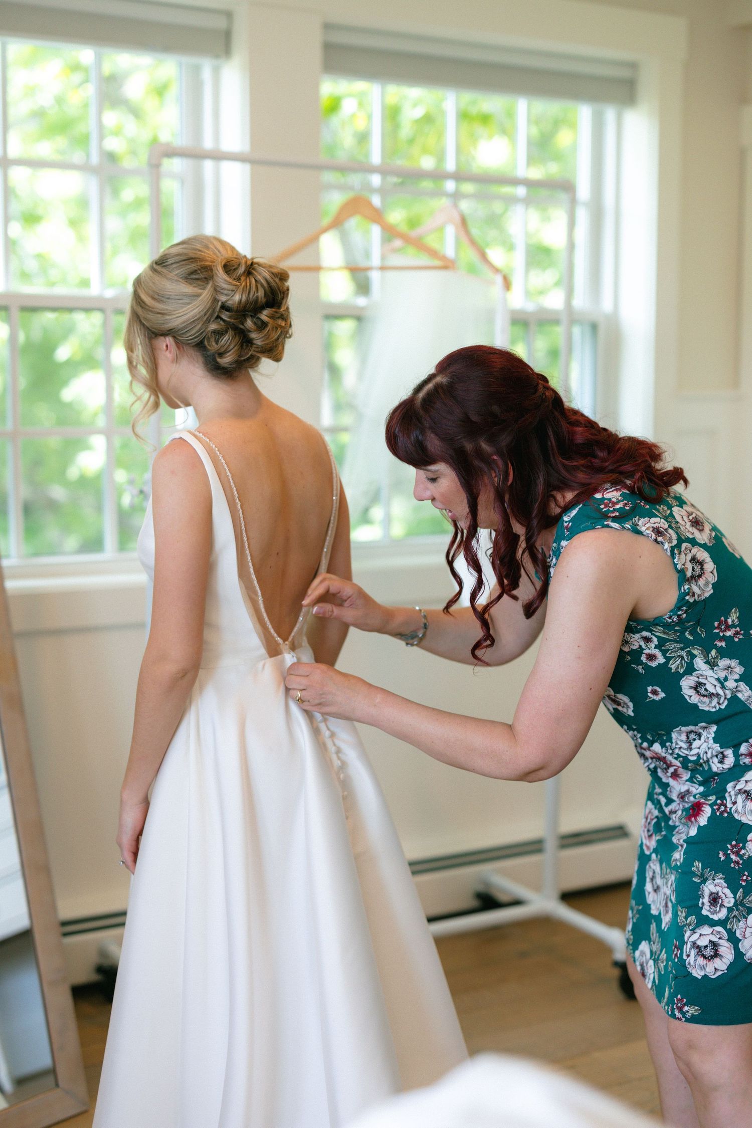 Bridesmaid helping bride button up elegant backless white wedding gown before ceremony.