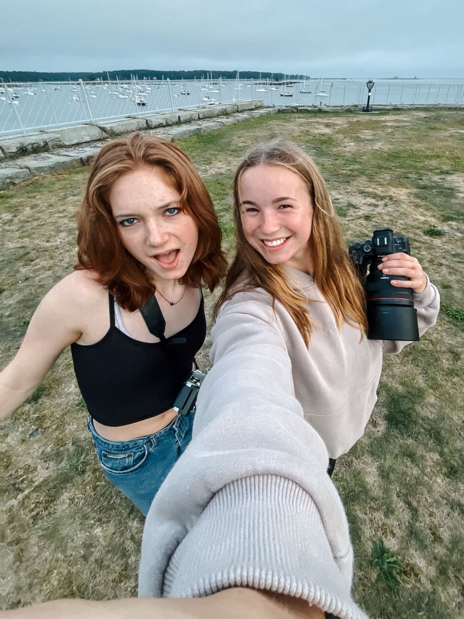 Two friends take a selfie on a grassy coastal overlook with boats and water visible in the background.
